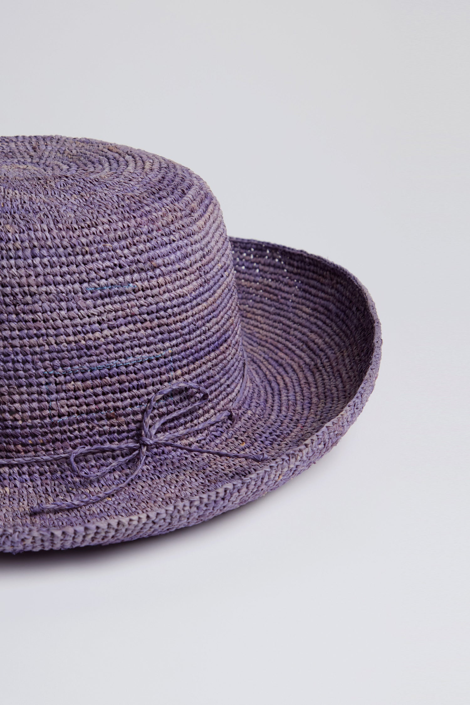 The Handwoven Raffia Hat from Rabarany, crafted with traditional techniques in Madagascar, showcases a lavender shade with a round crown and a small decorative bow on the brim.