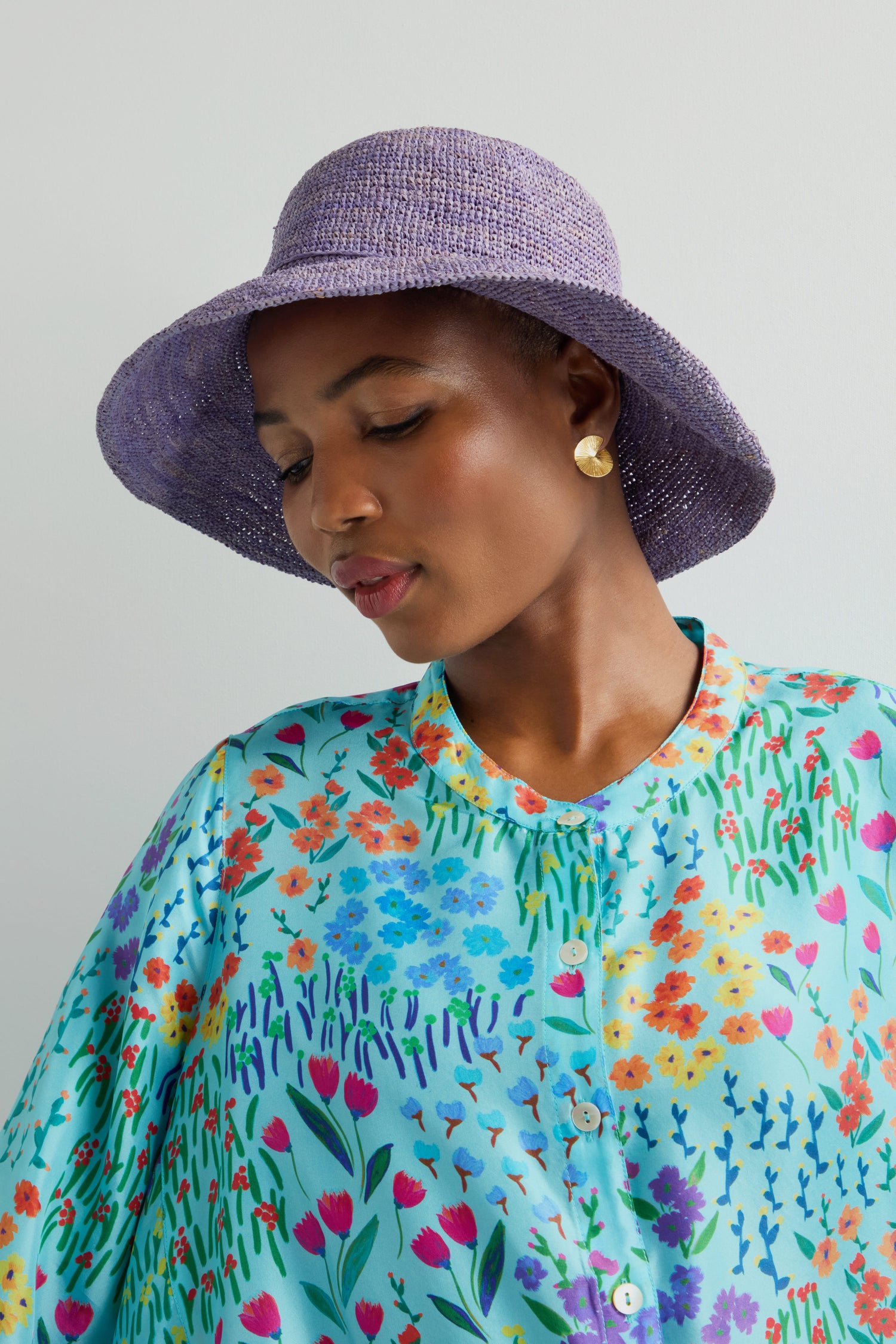 A person dons a Handwoven Raffia Hat in purple, crafted with Madagascar weaving, paired with a floral shirt and gold earrings, looking down against a plain backdrop.
