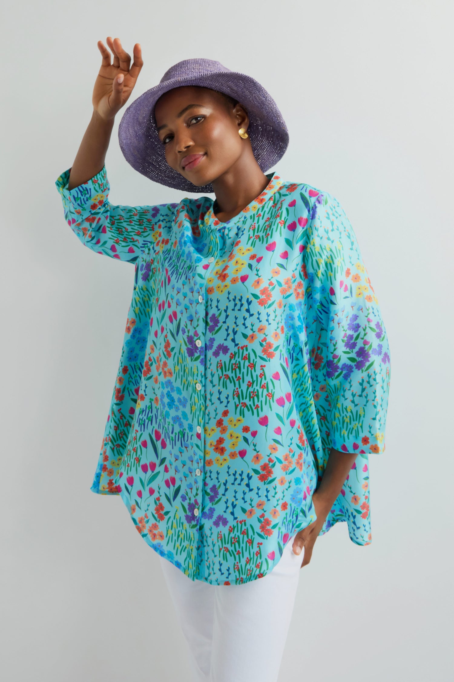 Person in a vibrant floral blouse and white pants, posing gracefully while touching their striking Handwoven Raffia Hat from Madagascar.