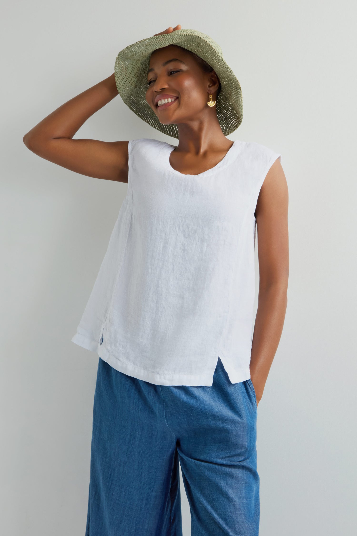 A person smiles wearing a Handwoven Raffia Hat showcasing Madagascar weaving techniques, paired with a sleeveless white top and blue pants. They pose against a plain background, one hand adjusting the breathable hat and the other in their pocket.