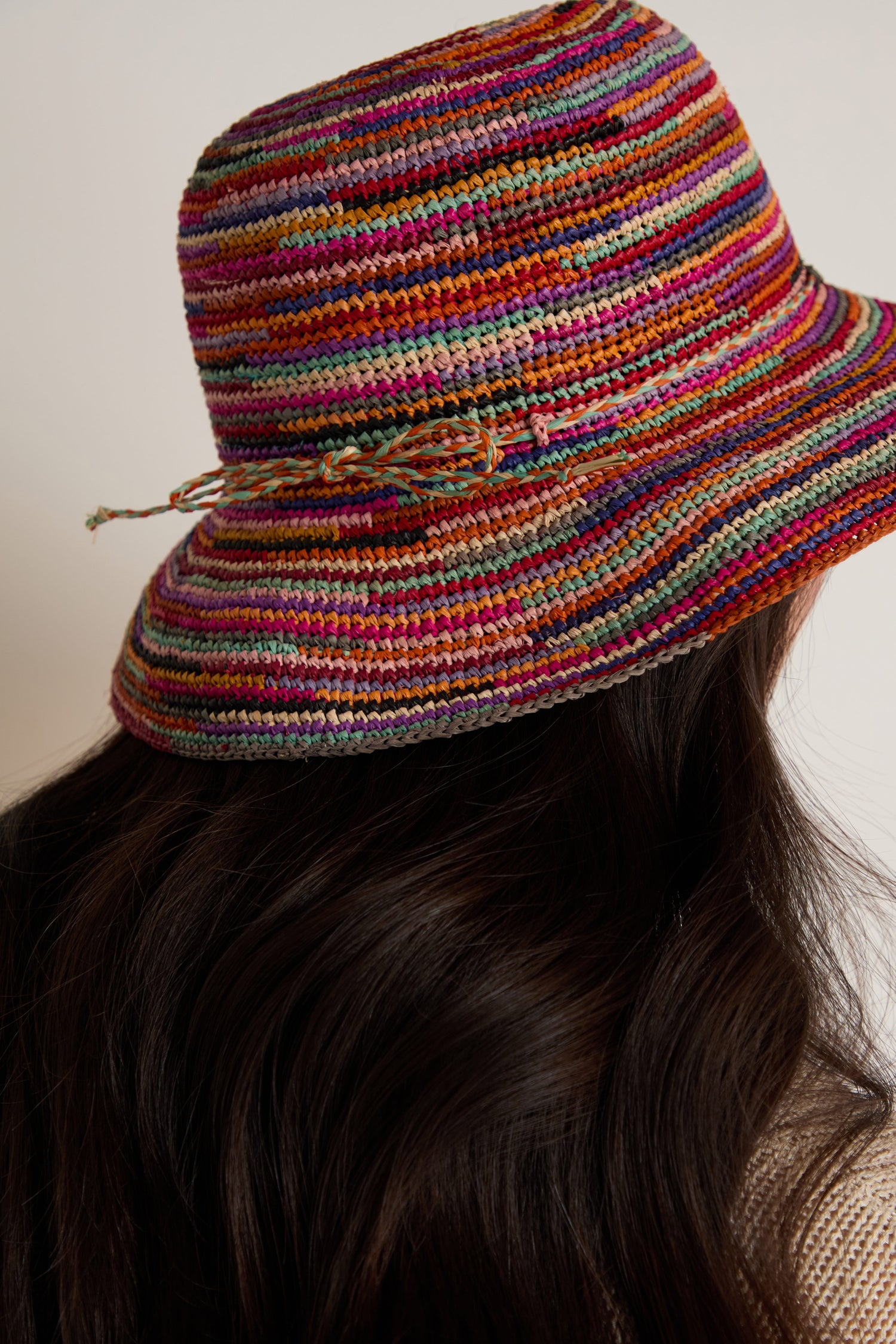 A person with long, dark hair is shown from behind wearing the Handwoven Rainbow Raffia Hat, featuring multicolored raffia and a small braided detail.