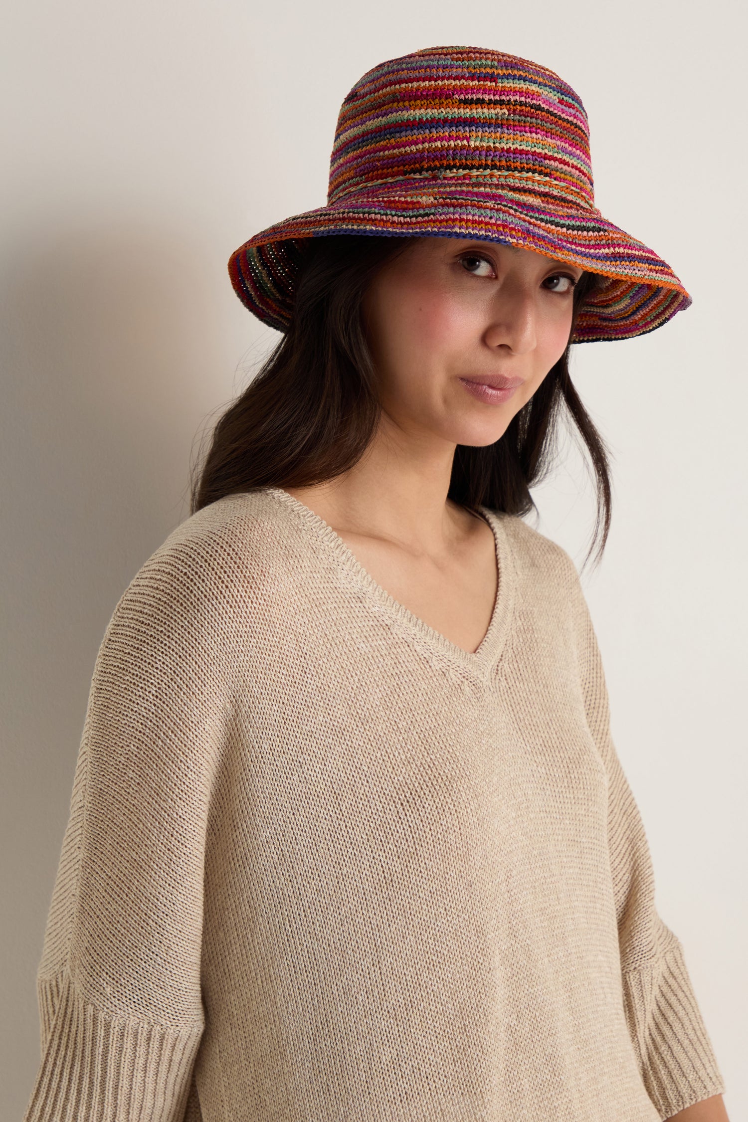 A woman in a beige knit sweater wears the Handwoven Rainbow Raffia Hat, standing against a plain light-colored wall.