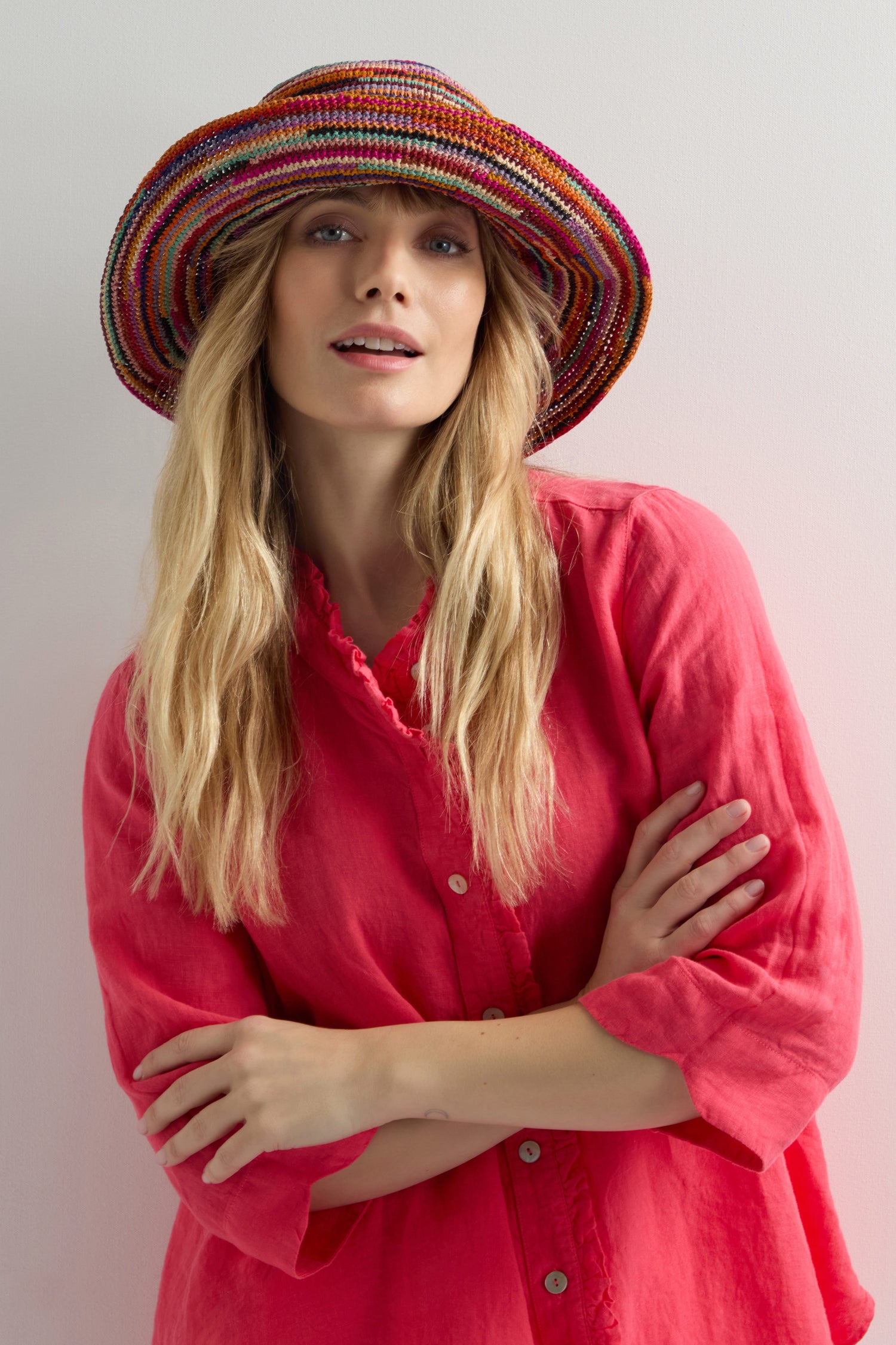A person with long blonde hair wears a Handwoven Rainbow Raffia Hat and a pink blouse, arms crossed, standing against a white background.