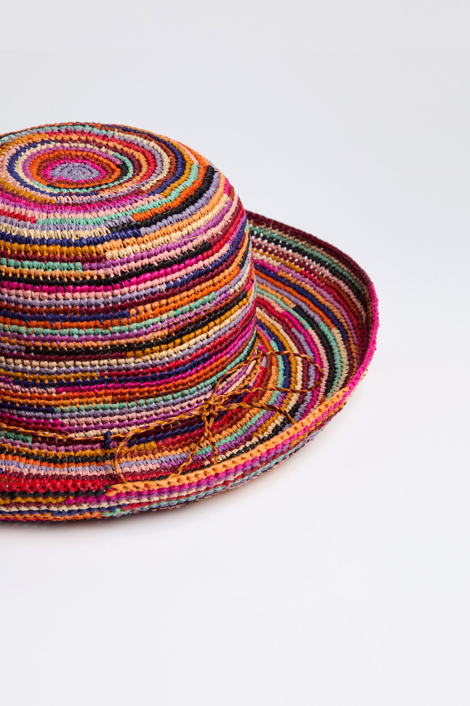 The Handwoven Rainbow Raffia Hat, with its vibrant concentric patterns, is beautifully displayed on a white surface, highlighting its craftsmanship and stylish sun protection.