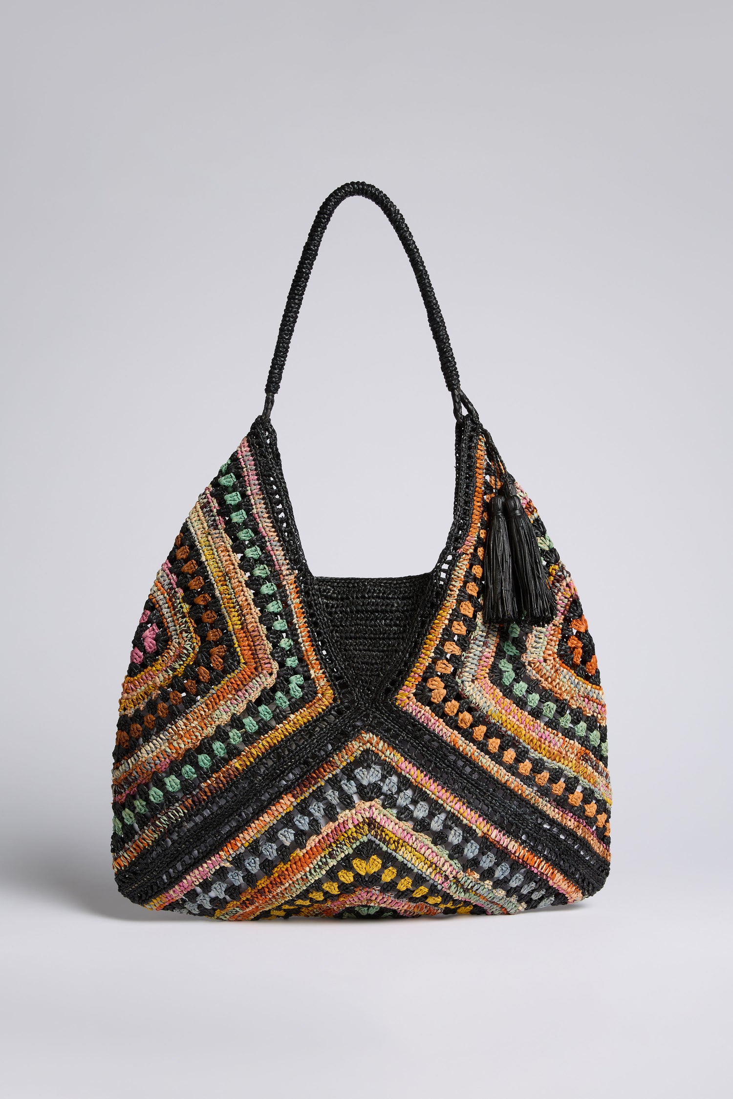 The Handwoven Bohemian Raffia Bag features a large, multicolored geometric patchwork design with a black handle and tassel, shown against a plain background.