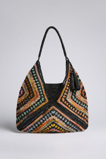 The Handwoven Bohemian Raffia Bag features a large, multicolored geometric patchwork design with a black handle and tassel, shown against a plain background.