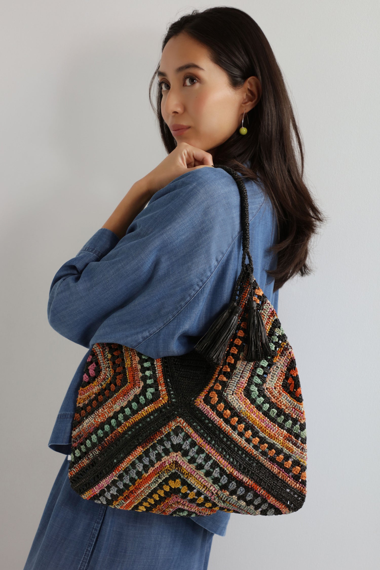 A woman in a blue outfit holds the Handwoven Bohemian Raffia Bag, featuring colorful geometric patchwork and tassels, against a plain light background.