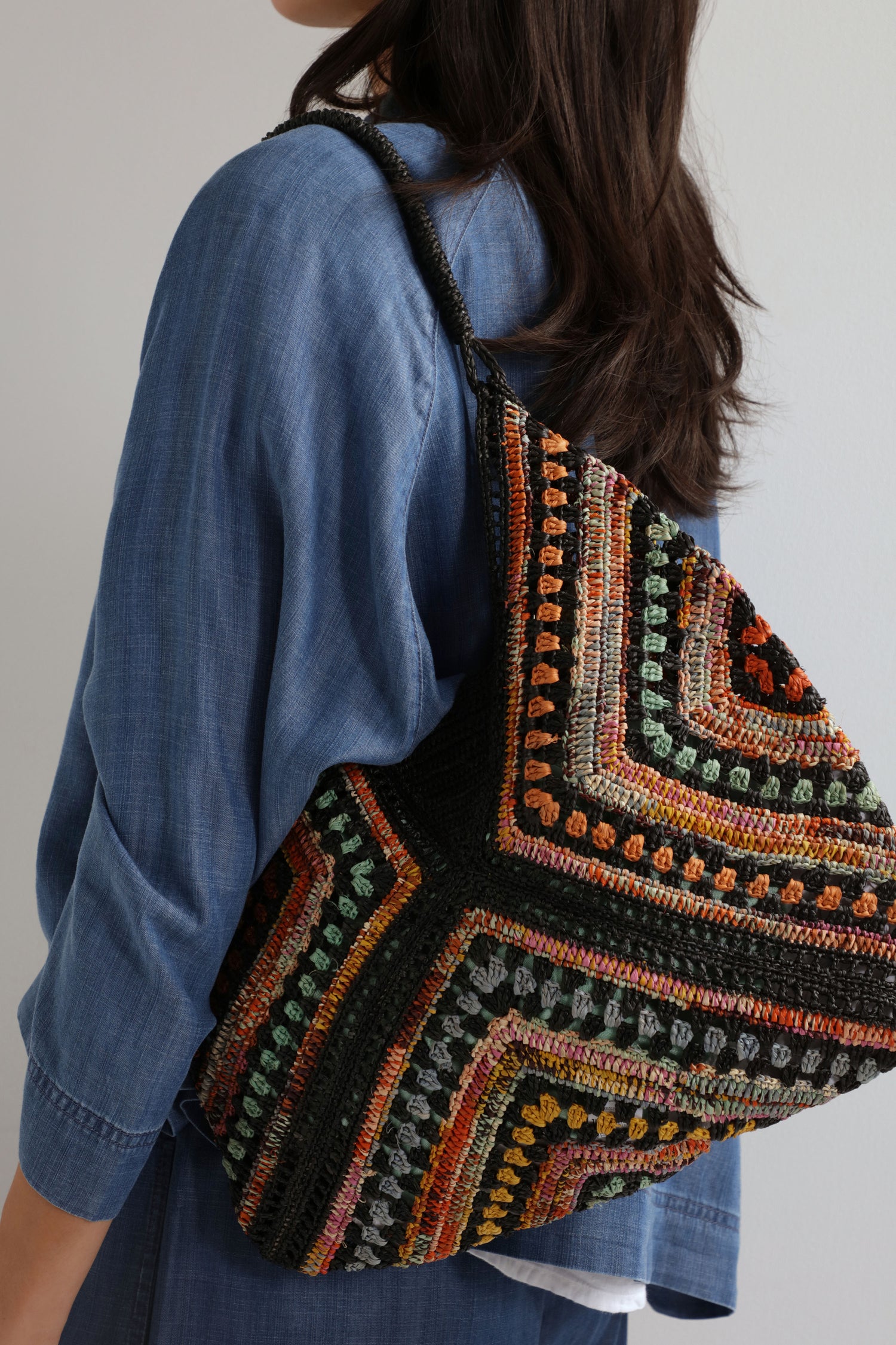 A person in a blue shirt carries a large Handwoven Bohemian Raffia Bag featuring colorful geometric patchwork and earthy tones.