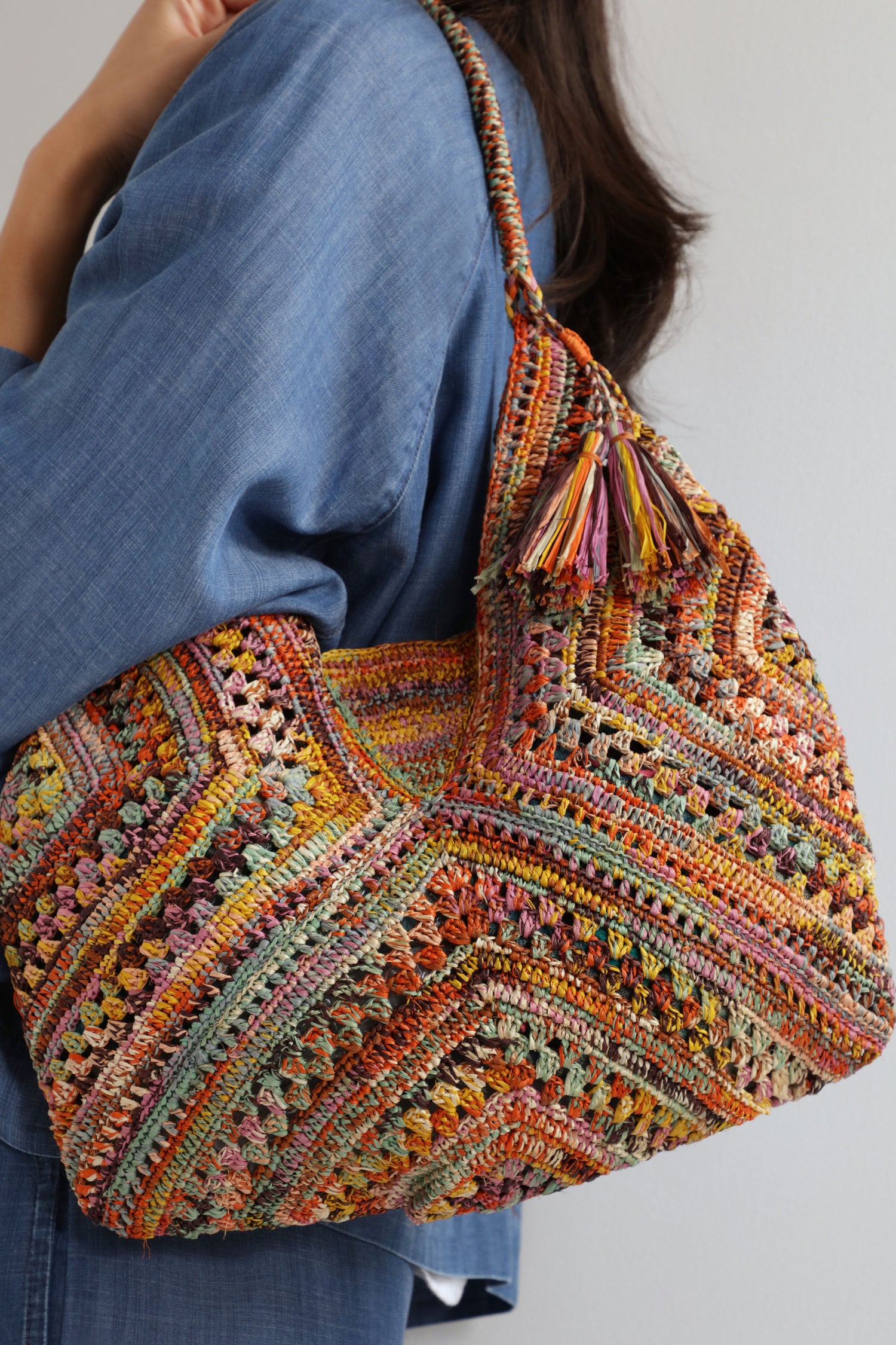 A person in a blue outfit carries the Handwoven Bohemian Raffia Bag, featuring a geometric patchwork design and a tassel detail on the handle.