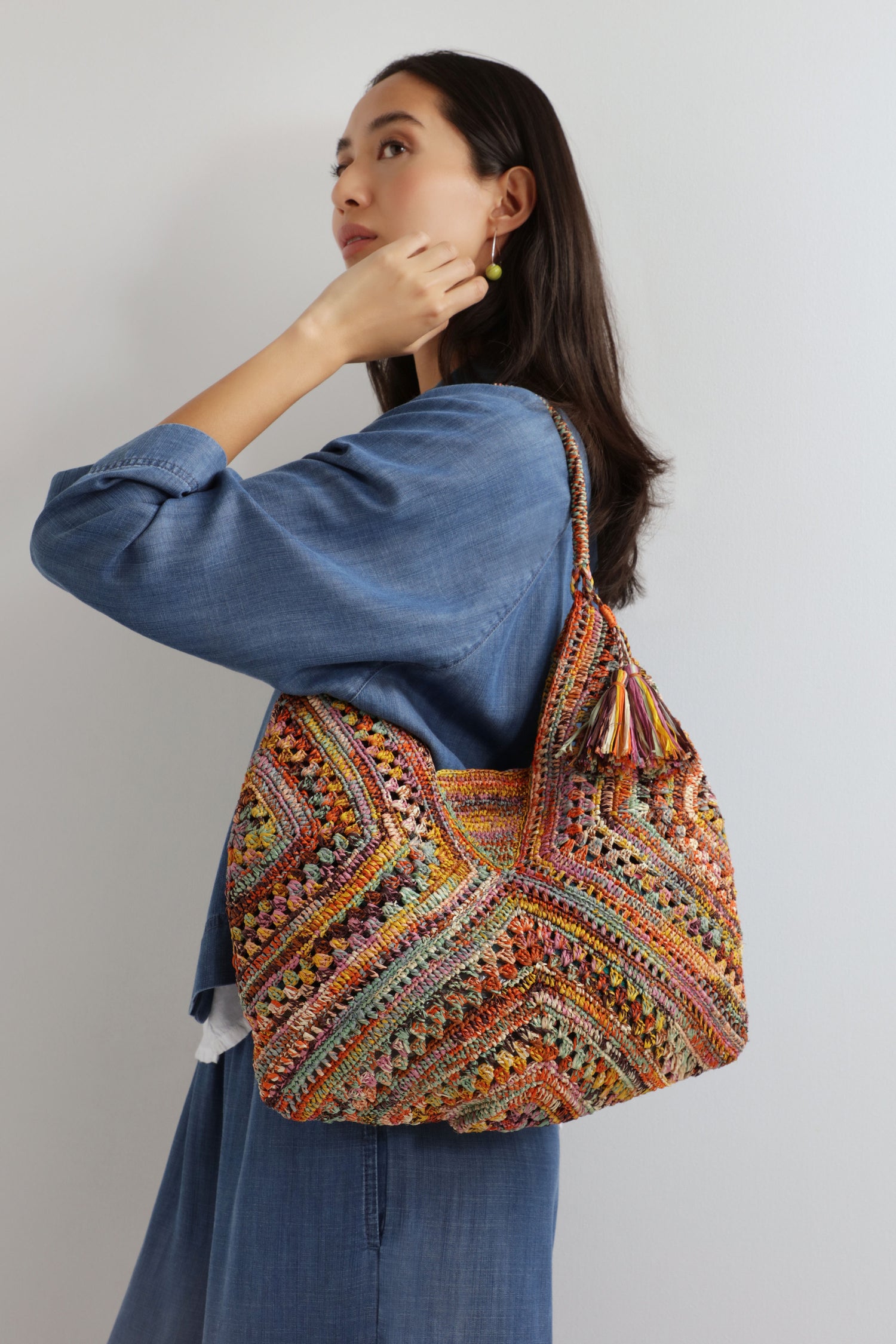 A woman in a blue denim outfit holds the Handwoven Bohemian Raffia Bag, which features a large, colorful geometric patchwork design and tassel details, standing against a plain background.