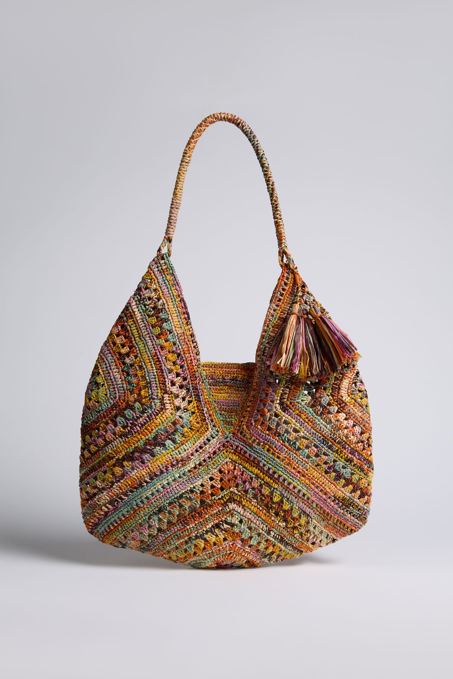 The Handwoven Bohemian Raffia Bag showcases a multicolored geometric patchwork, a single handle, and a decorative tassel, displayed against a plain light background.