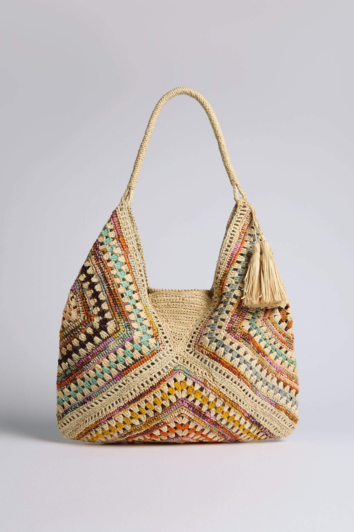 The Natural Handwoven Bohemian Raffia Bag is multicolored with a large handle and tassel, showcasing a geometric patchwork design and a soft, relaxed shape.