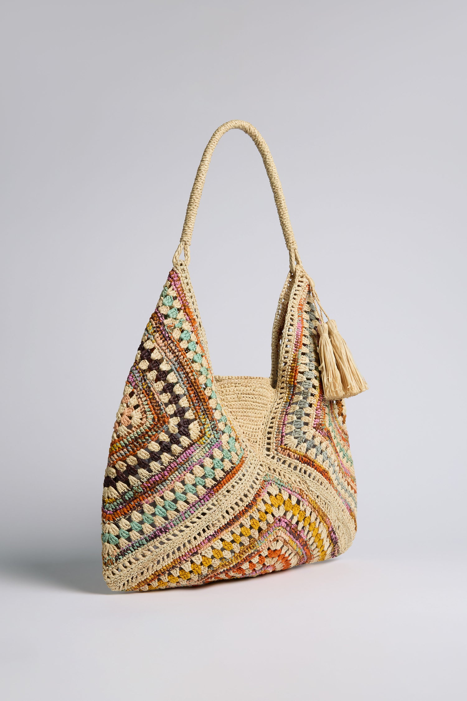 The Natural Handwoven Bohemian Raffia Bag features a multicolored crochet pattern, thick handle, tassel accent, and subtle geometric patchwork design, displayed against a plain backdrop.