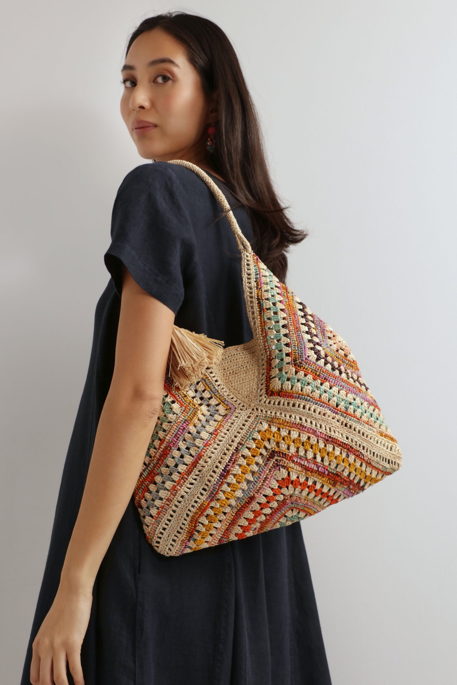 A woman in a dark dress stands sideways, looking over her shoulder, carrying the Natural Handwoven Bohemian Raffia Bag with a bold geometric patchwork design.