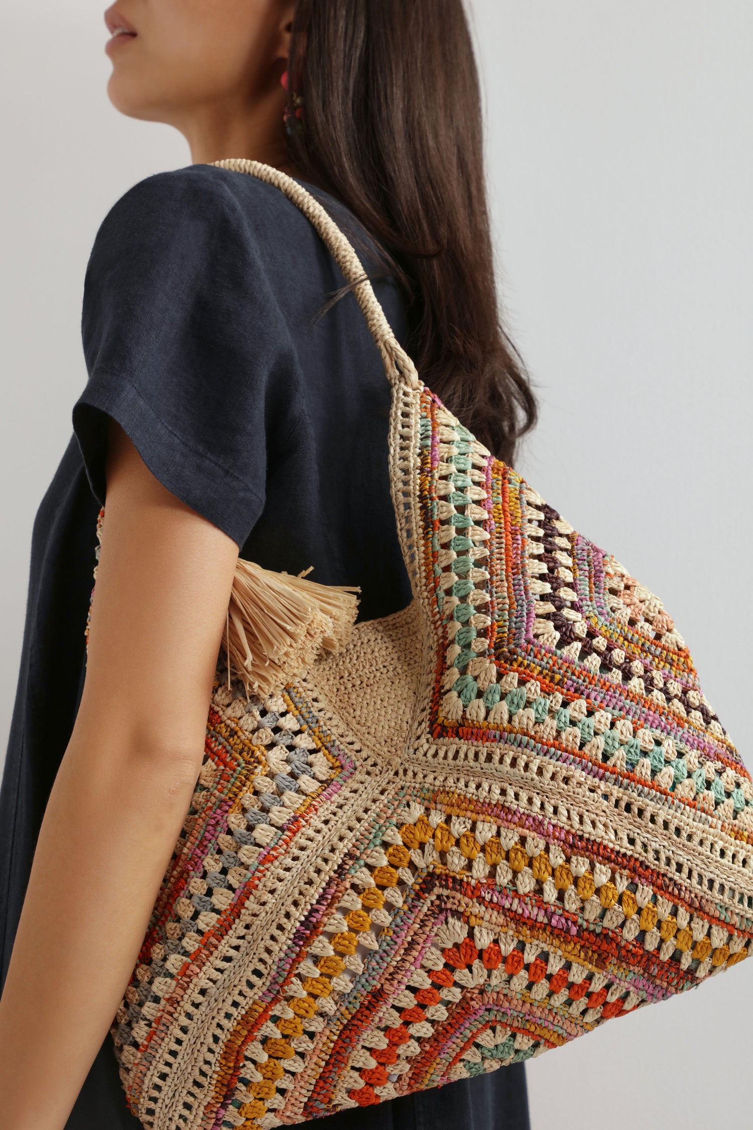 A woman in a dark short-sleeve top carries the Natural Handwoven Bohemian Raffia Bag with a geometric patchwork design and tassel detail over her shoulder.