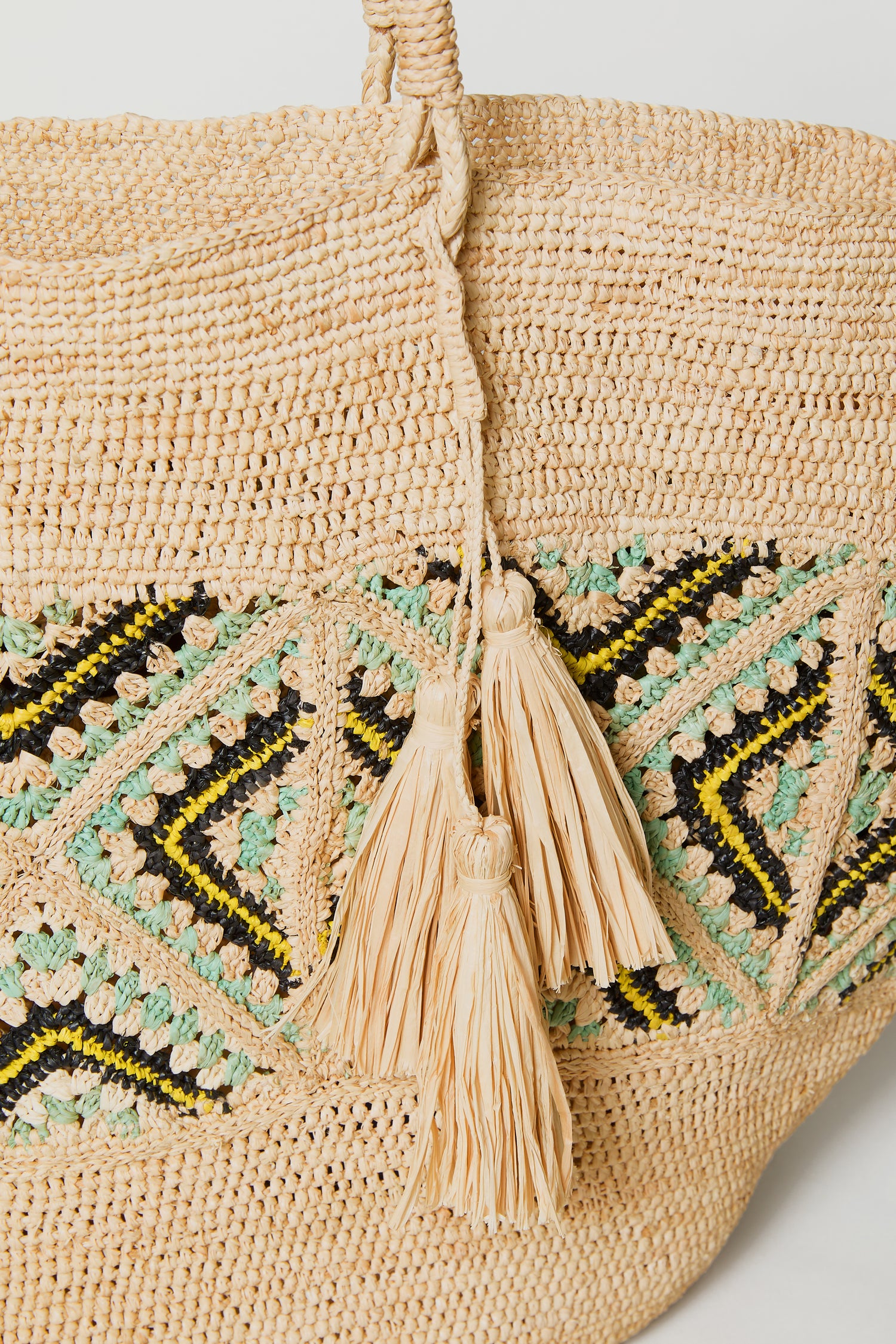 Close-up of the Handwoven Raffia Mosaic Bag, showcasing geometric patterns in black, yellow, and green, and two braided tassels on the handle.