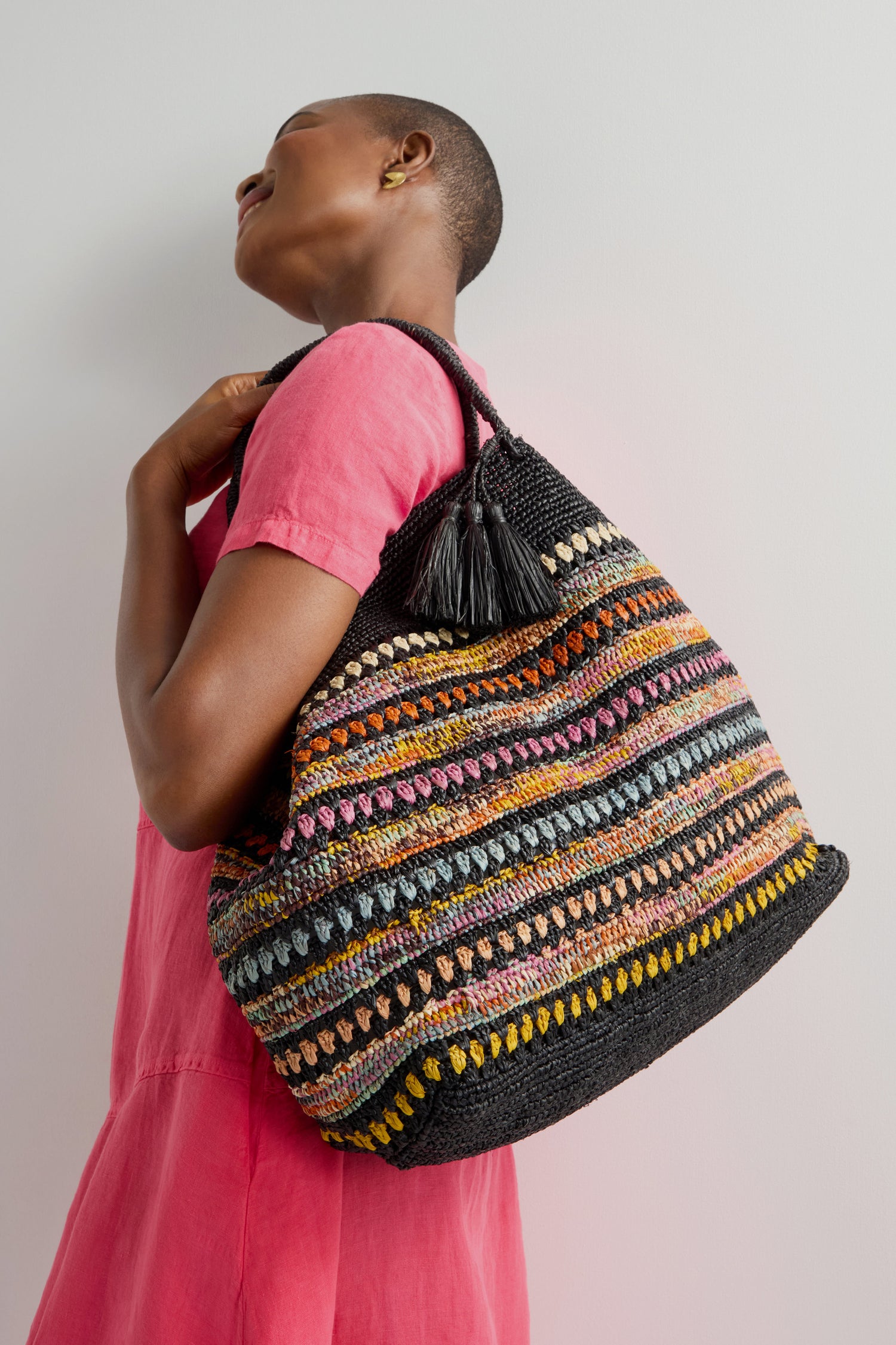 A person in a pink dress holds the Handwoven Crochet Stripe Raffia Bag, featuring a colorful design with a black base and tassel, reflecting artisanal craftsmanship against a plain background.