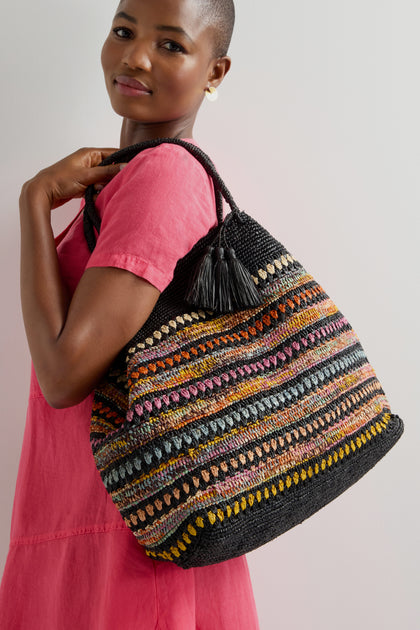 In a pink dress, a person shows off the exquisite artisanal craftsmanship of a large Handwoven Crochet Stripe Raffia Bag, featuring colorful stripes and tassel details.