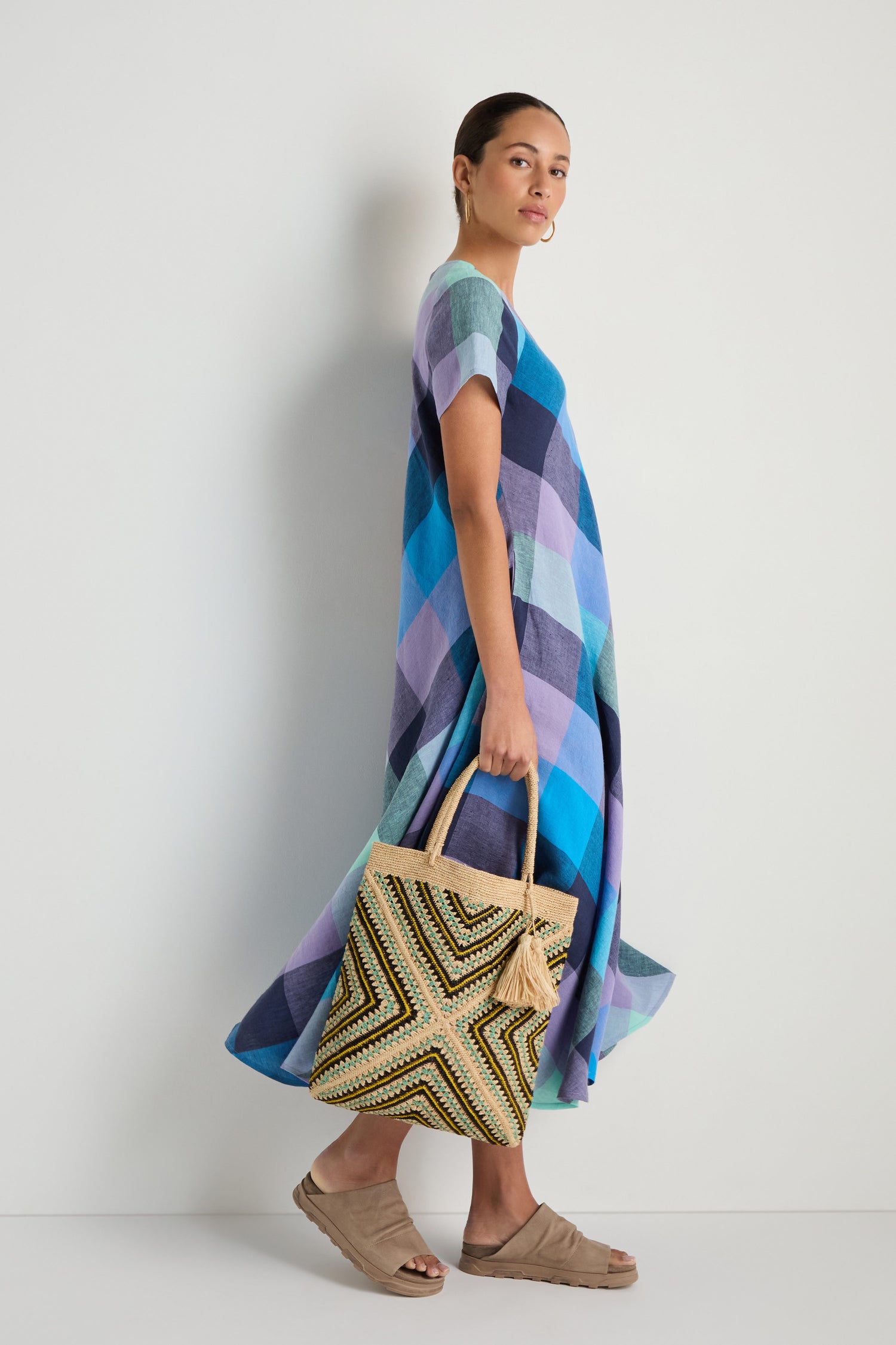 A woman in a blue and purple checkered dress stands against a white background, holding a Handwoven Raffia Tote Bag and wearing beige sandals.