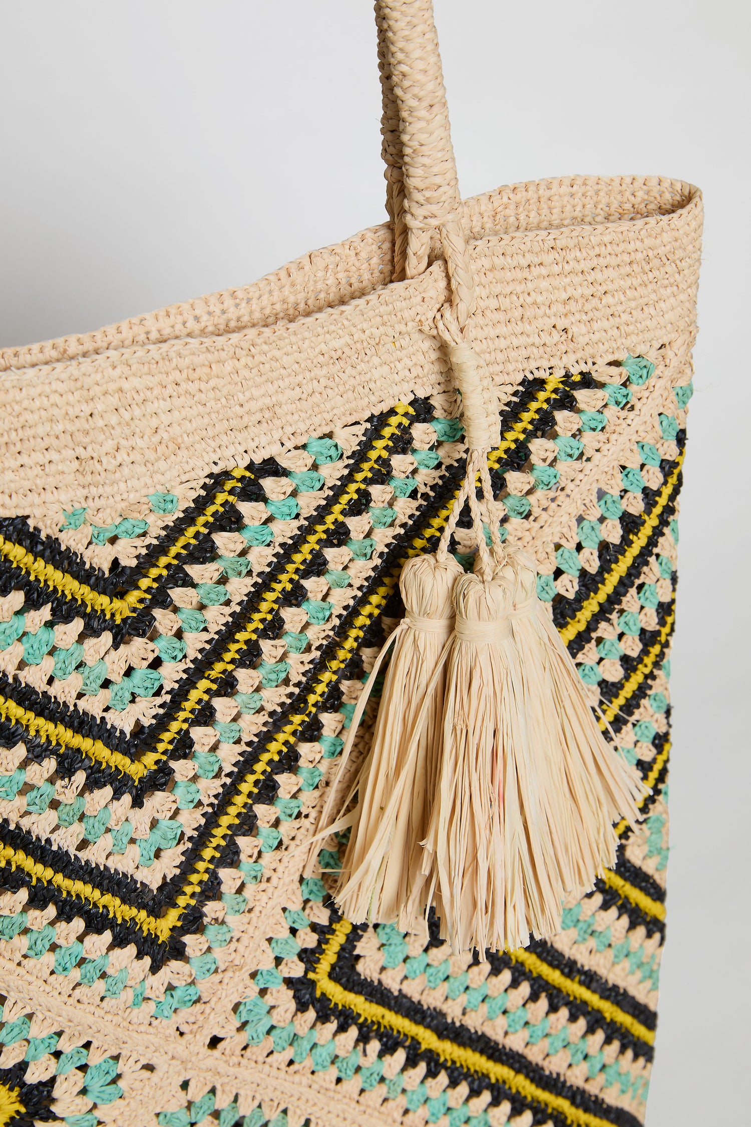 Close-up of the Handwoven Raffia Tote Bag, crafted in Madagascar with black, yellow, and turquoise geometric patterns and two tassels on the handle.