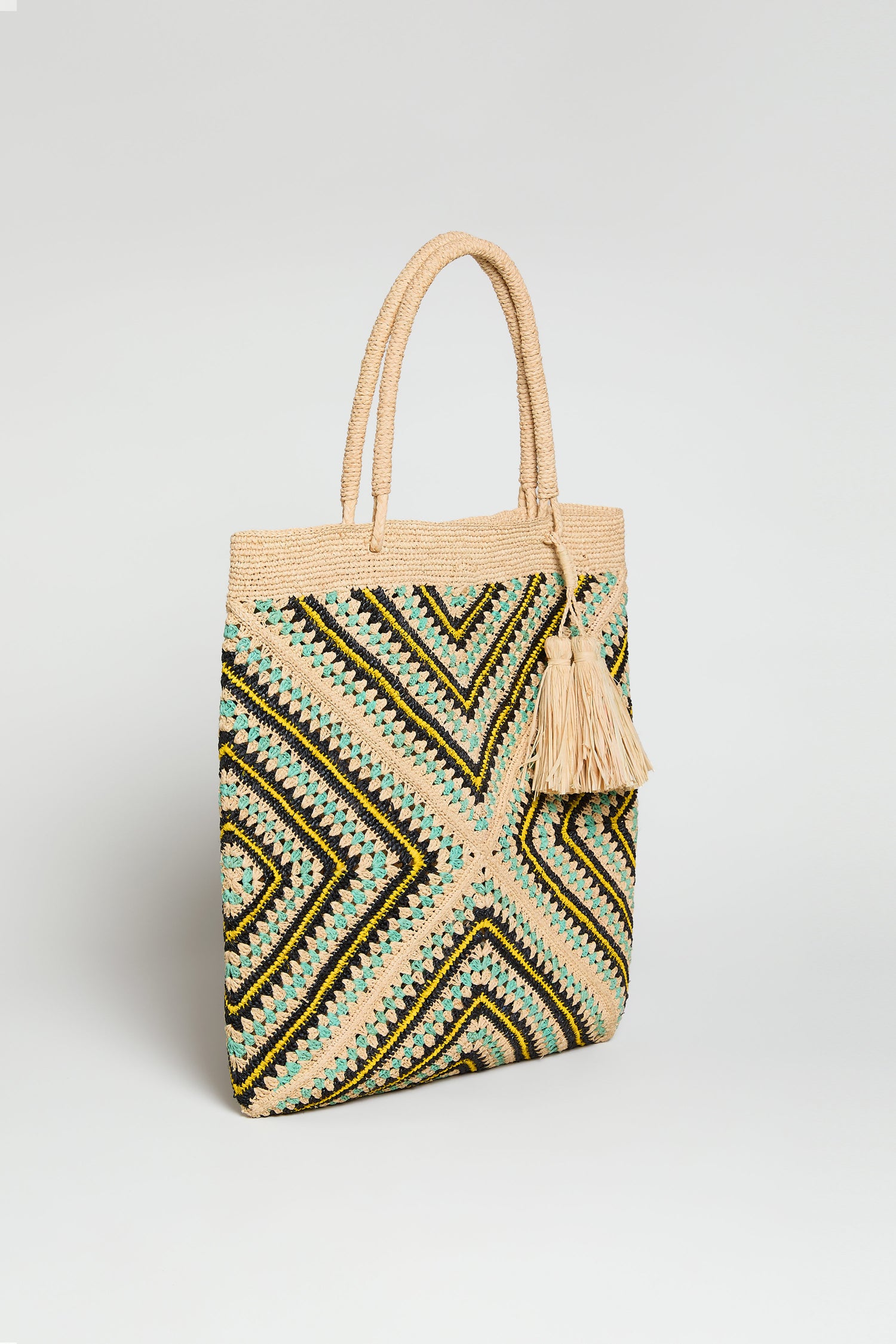 The Handwoven Raffia Tote Bag features artisan craftsmanship, geometric black, yellow, and green patterns, two handles, and a large beige tassel.