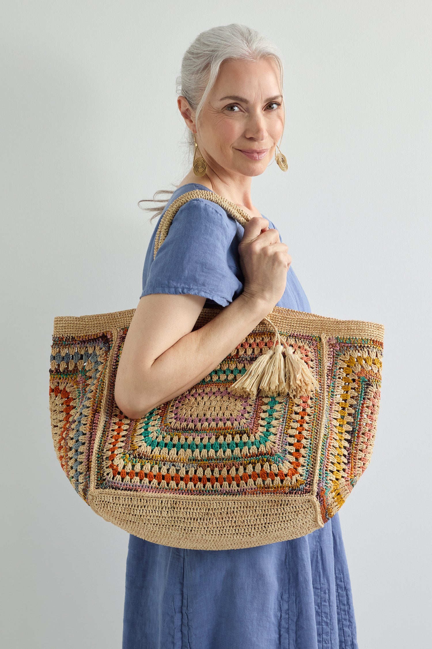 A woman with gray hair in a blue dress stands against a plain light background, holding the Handwoven Crochet Raffia Bag accented with tassels.