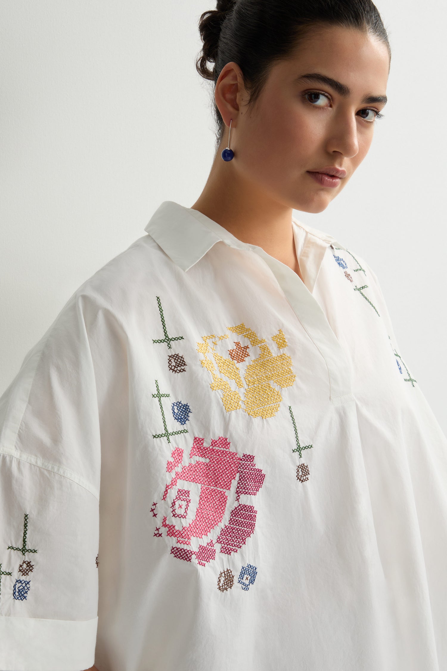 An individual wears the Cross Stitch Embroidery Tunic, a white shirt featuring an abstract floral pattern, while looking toward the camera.