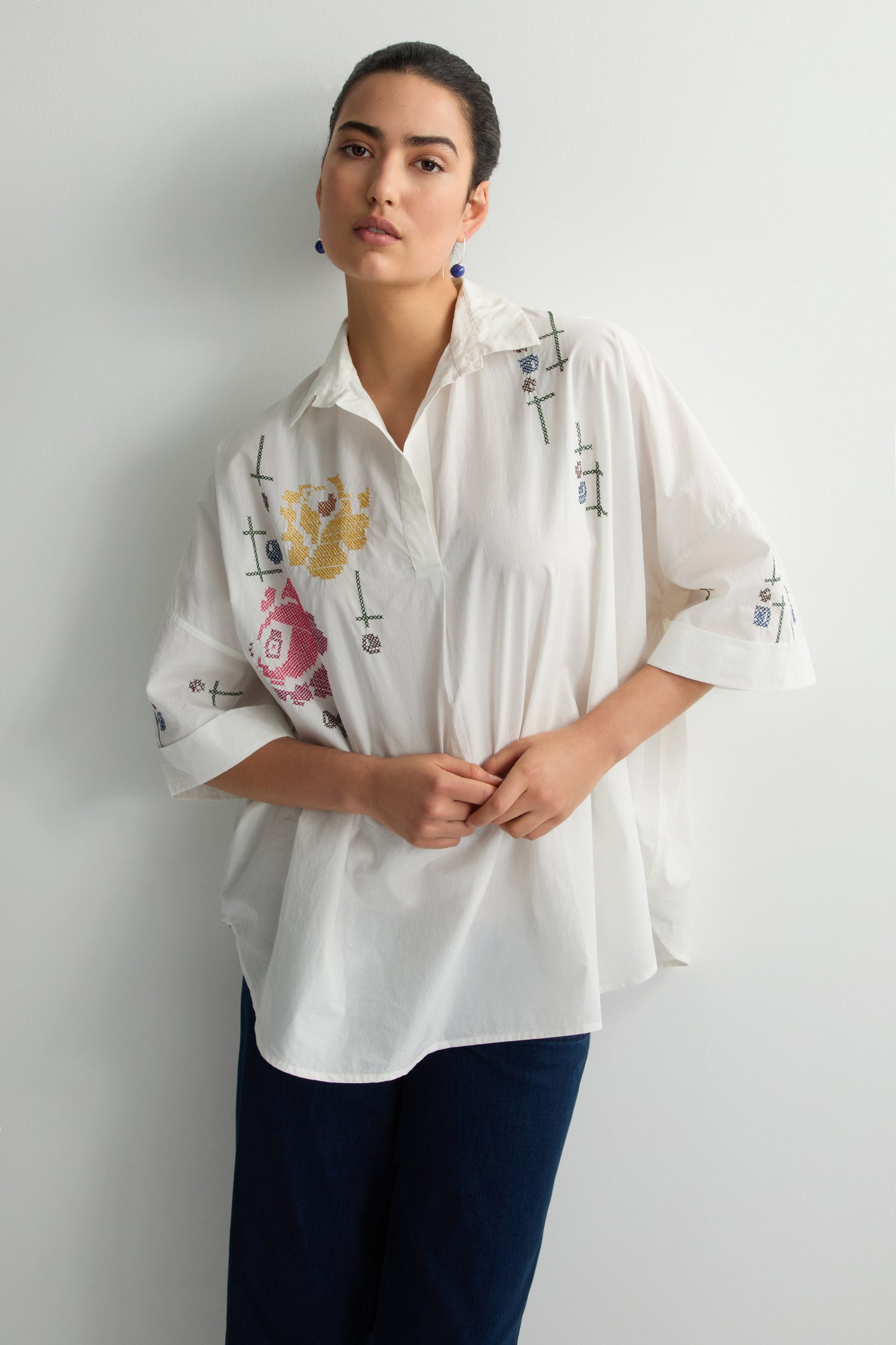 A person in a Cross Stitch Embroidery Tunic and dark pants stands against a plain white background, exuding subtle charm through the tunic's intricate floral motifs.