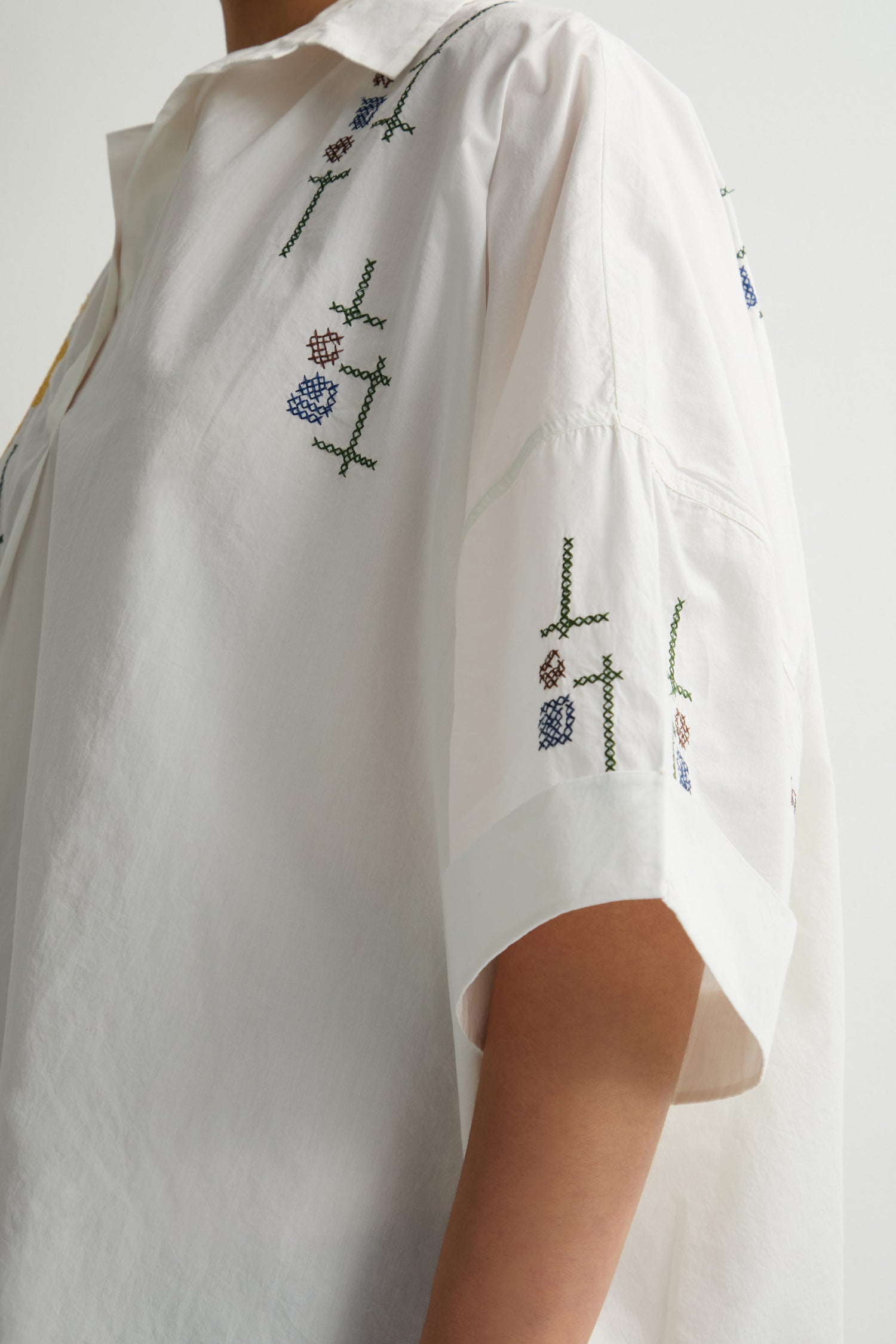 A person wearing a Cross Stitch Embroidery Tunic featuring a loose-fitting white design with an abstract floral pattern and exquisite cross-stitch embroidery on the shoulder and sleeve.