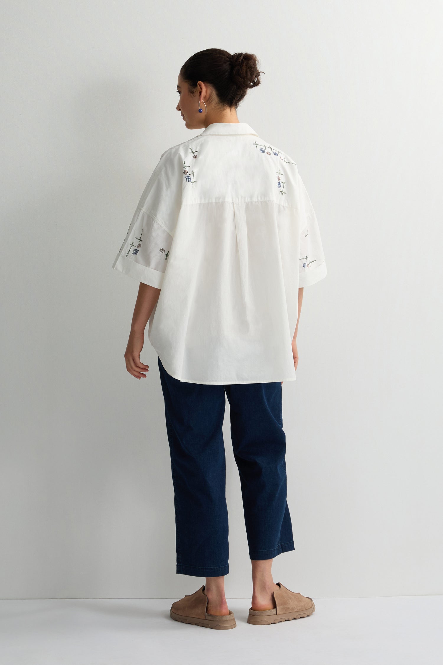 Facing away from the camera, a person stands against a white wall, wearing a Cross Stitch Embroidery Tunic with dark blue pants and brown shoes, hair styled in a bun.