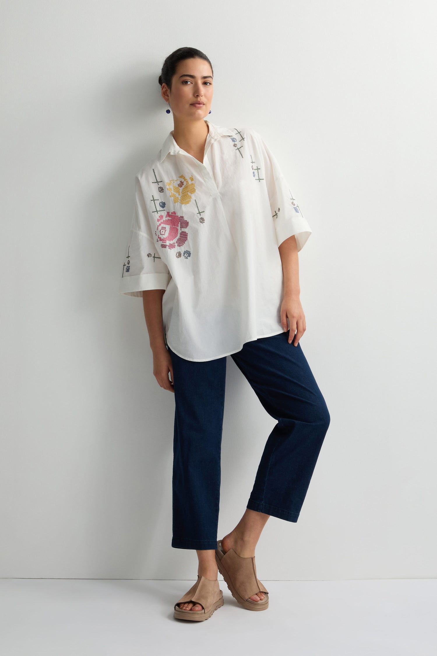 A person stands against a white wall wearing a Cross Stitch Embroidery Tunic, dark blue pants, and beige sandals.
