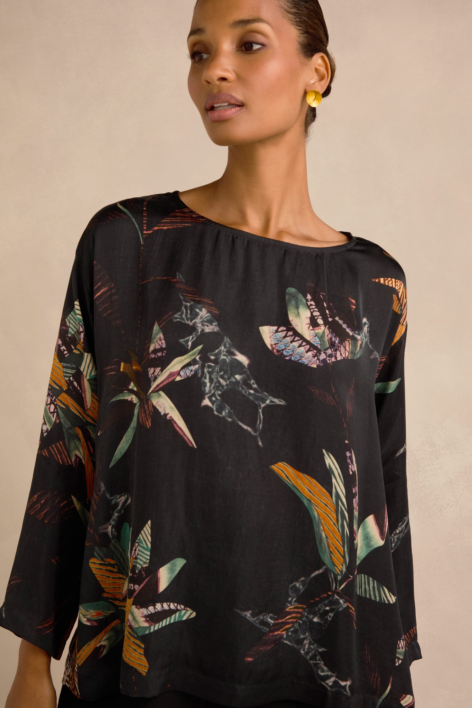 A woman is wearing the Abstract Floral Boxy Top featuring colorful patterns, paired with small yellow earrings, standing against a neutral background.