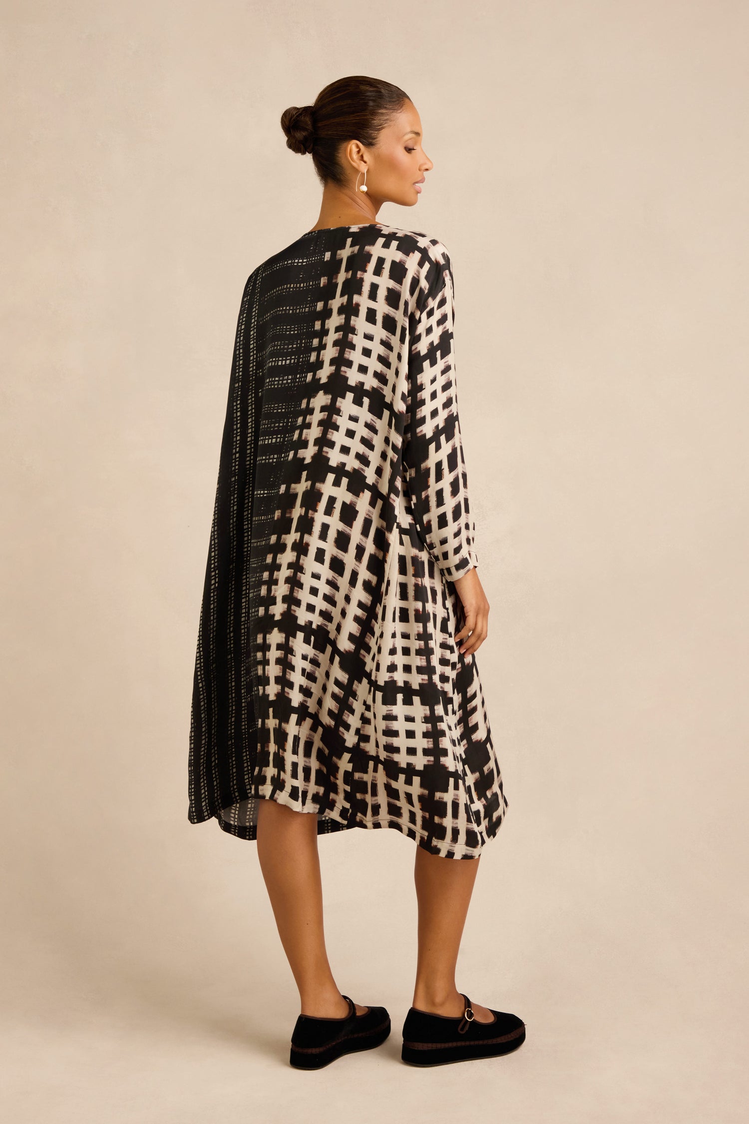 A woman stands facing away, wearing the Check and Dash Easy Dress—a monochrome piece with a black and white plaid pattern and A-line silhouette—paired with black shoes and her hair in a bun, set against a plain beige background.