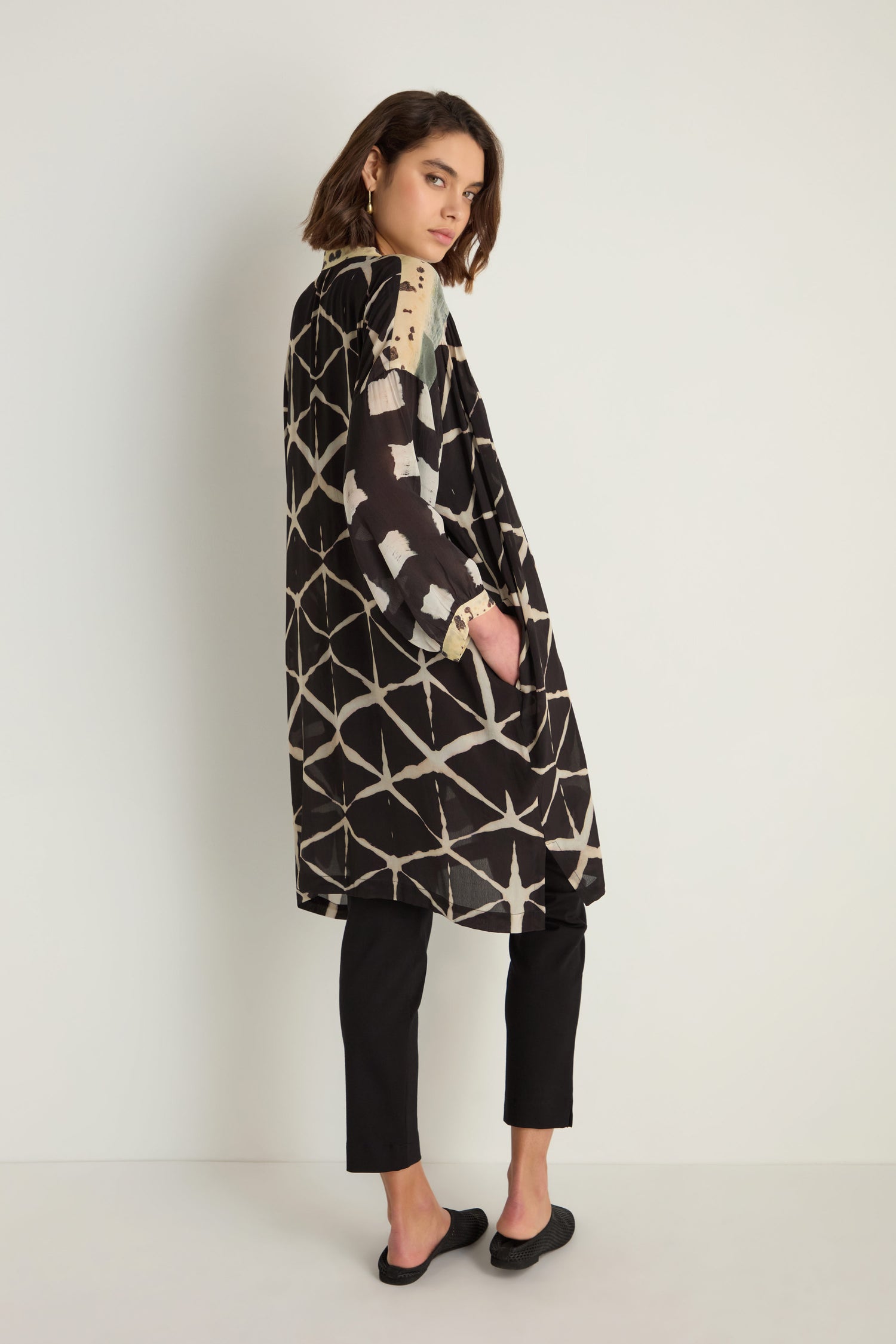 A person stands sideways with one hand in their pocket, wearing a relaxed fit, black and beige Shibori Print Long Shirt over black pants against a plain light background.