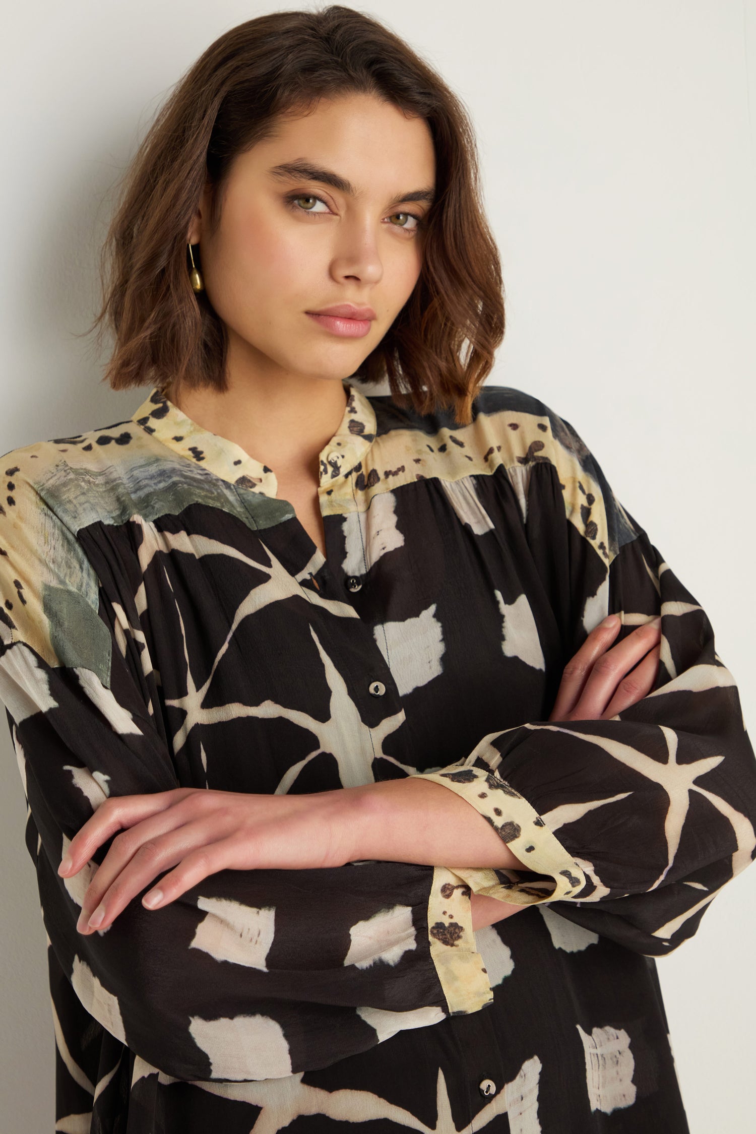 A woman with short brown hair models the Shibori Print Long Shirt in a relaxed fit, standing against a white wall with her arms crossed and looking at the camera.