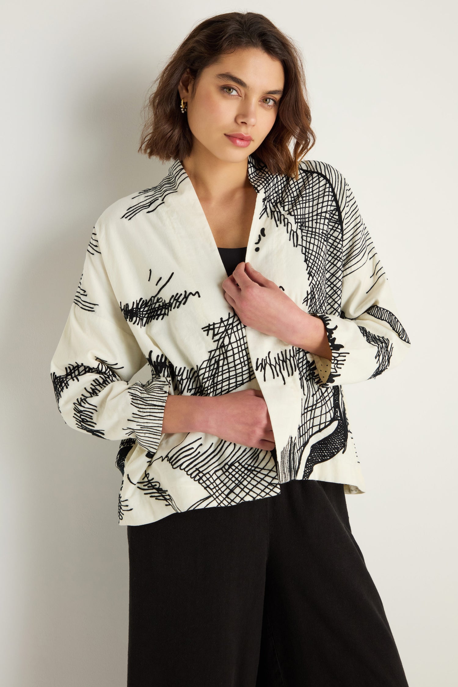 A woman stands against a light background wearing the Embroidered Sketch Jacket—white cotton with black abstract patterns—and black pants, effortlessly showcasing artisan design while looking at the camera.