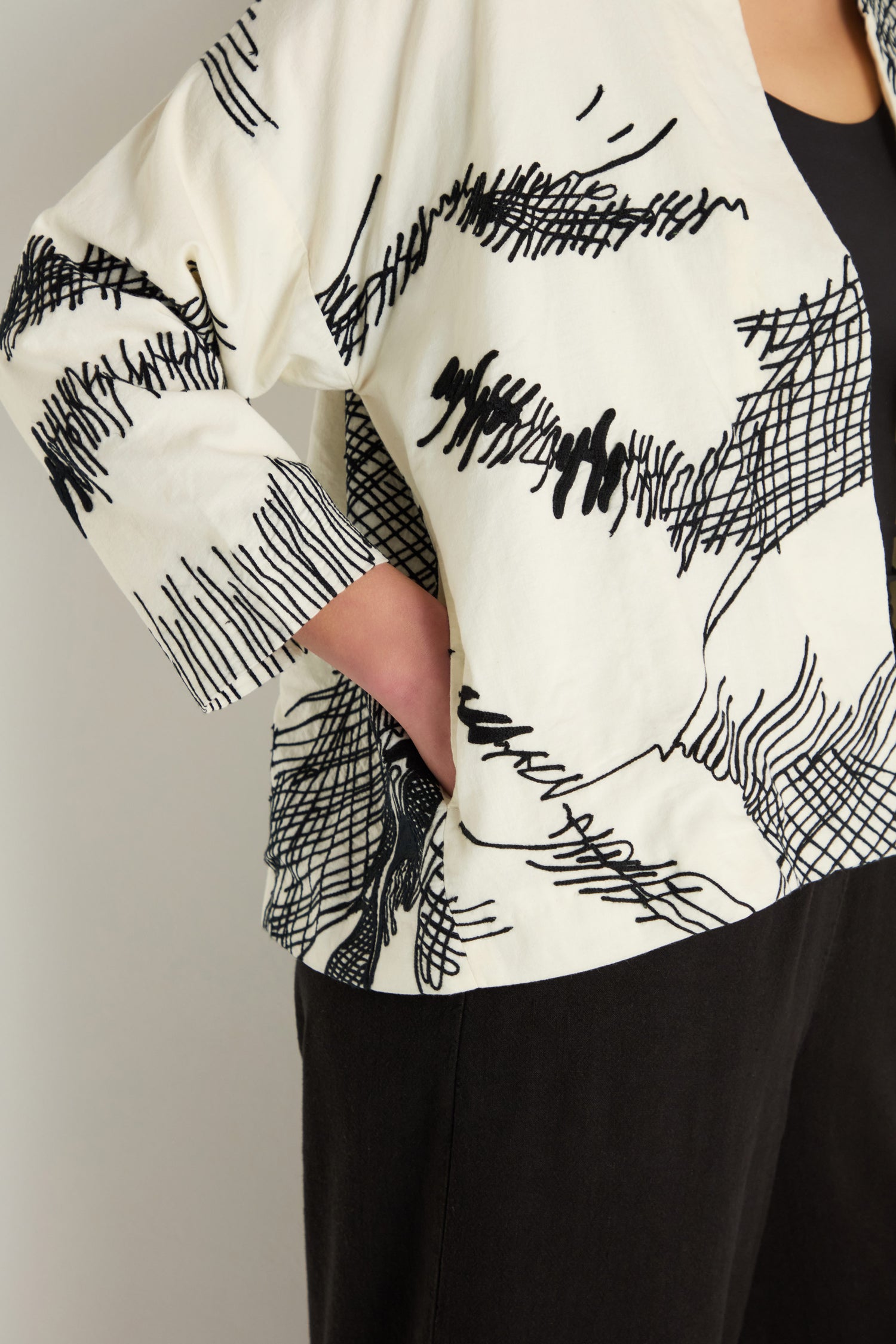 A person stands with one hand on their hip, wearing the Embroidered Sketch Jacket—an artisan-designed white cotton jacket featuring black abstract line patterns—and black pants.
