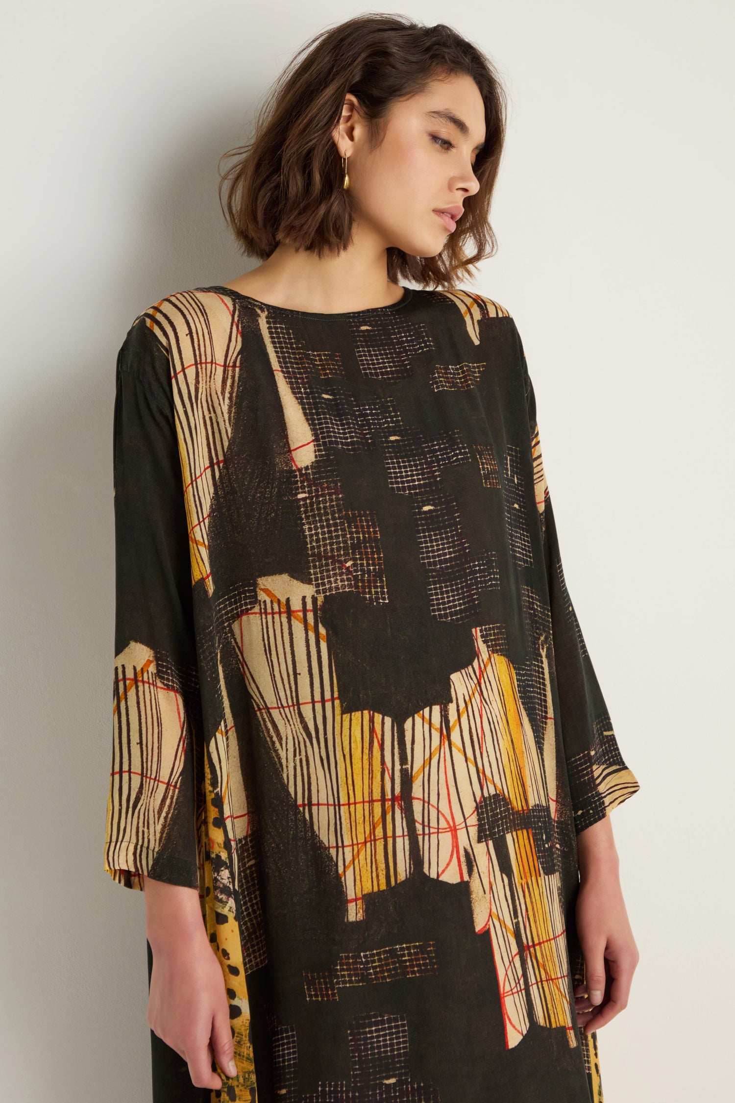 A woman with shoulder-length brown hair wears the Klimt Print Easy Dress, featuring painterly black, beige, and yellow prints, as she stands against a plain wall.