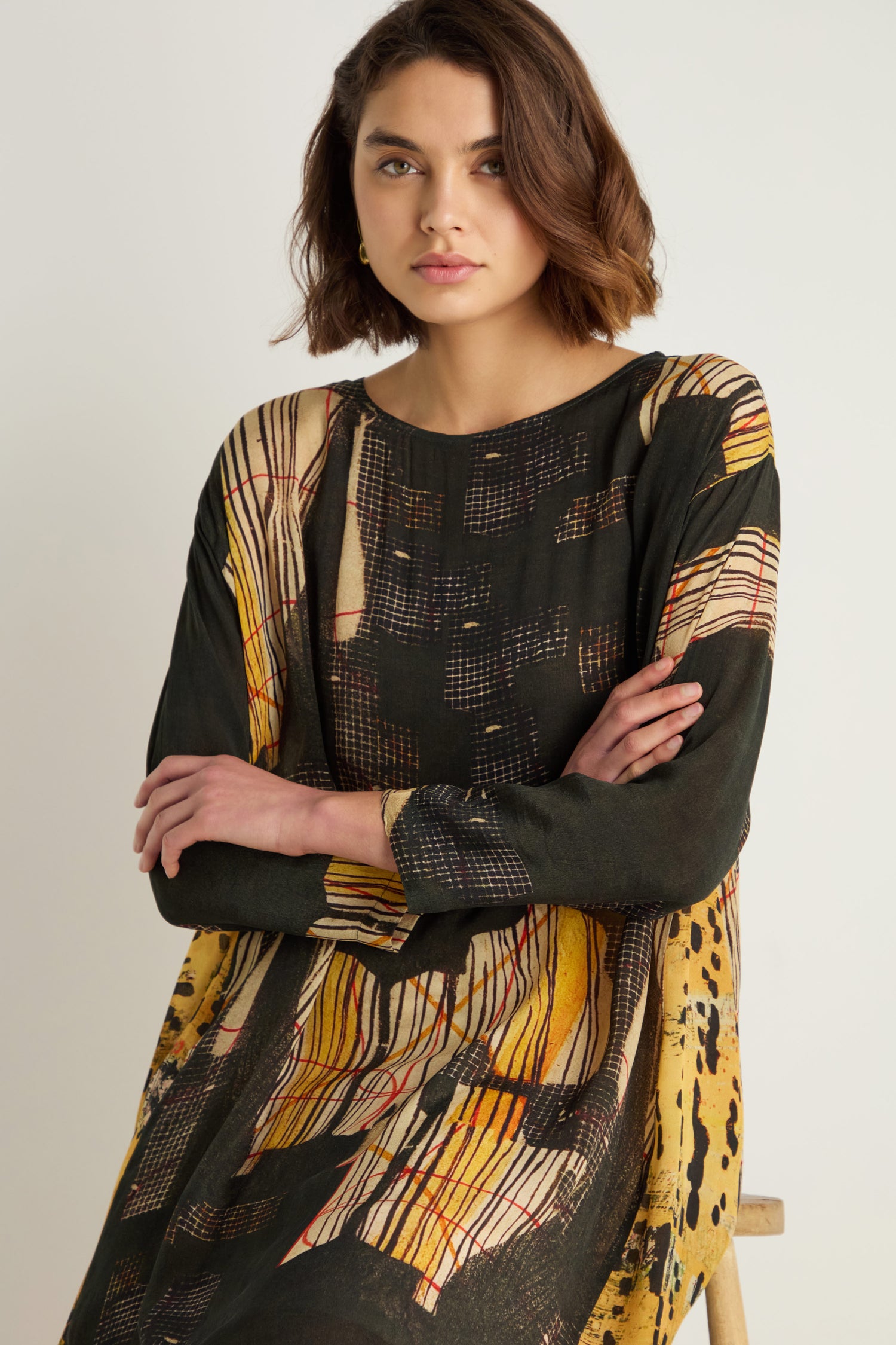 A woman with short brown hair sits with arms crossed, wearing a long-sleeved Klimt Print Easy Dress in painterly yellow, black, and red on a soft cupro-viscose blend.