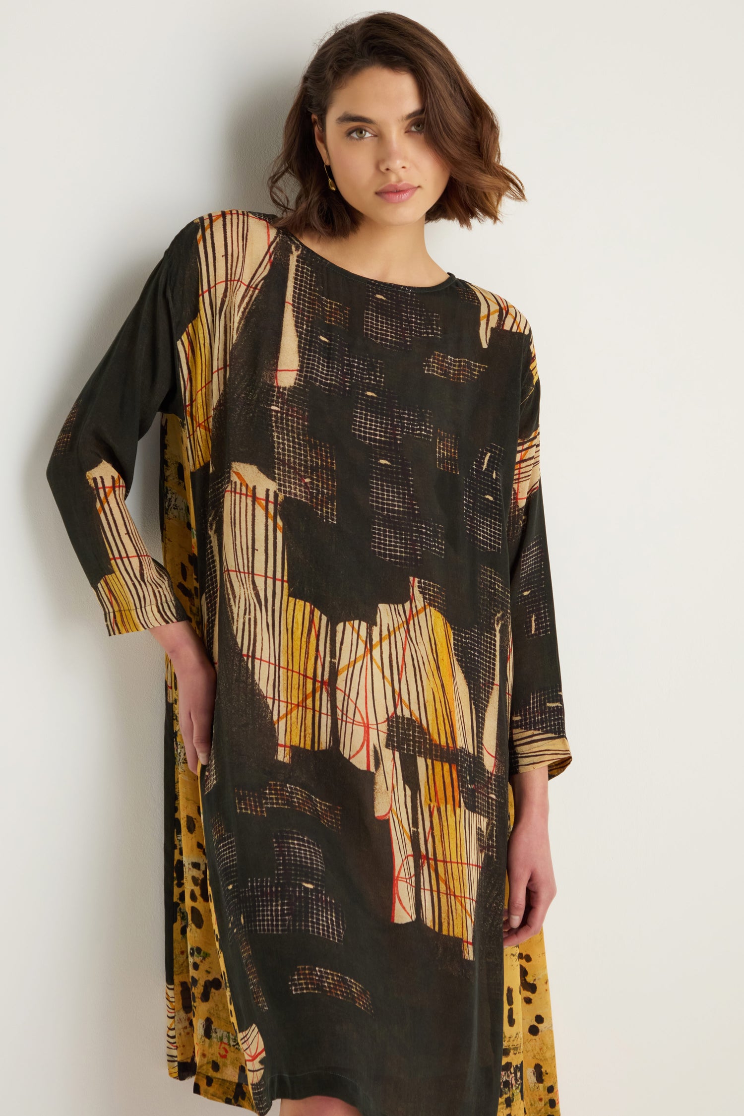 A woman wears the Klimt Print Easy Dress, made from a soft cupro viscose blend with painterly black, yellow, and beige prints, standing against a plain light-colored wall.