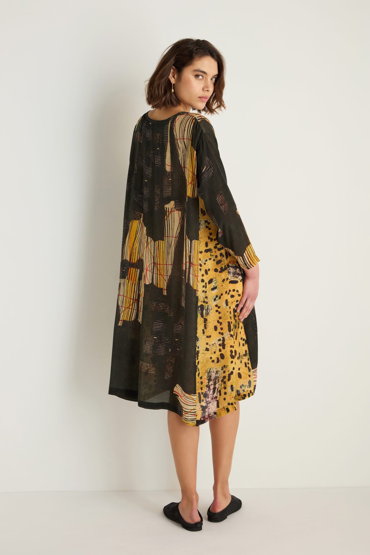 A woman stands against a white wall in the Klimt Print Easy Dress, a loose, long-sleeved style with black and yellow abstract prints, crafted from a soft cupro-viscose blend and paired with black flat shoes.