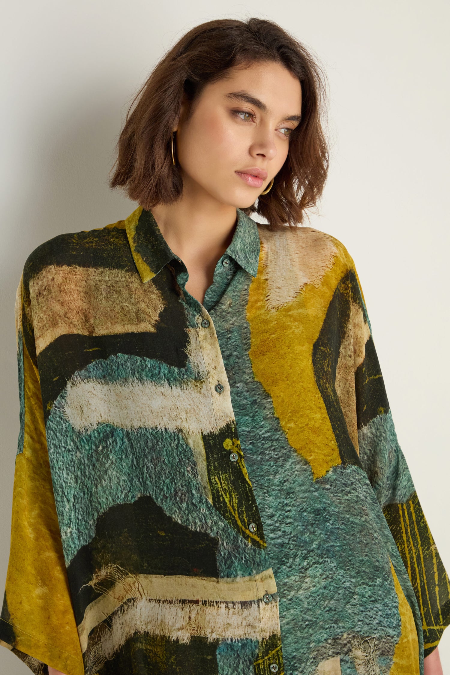 A woman with short brown hair wears the Ochre Print Shirt Dress, featuring loose painterly prints in abstract green, yellow, and tan patterns, standing against a plain white background.
