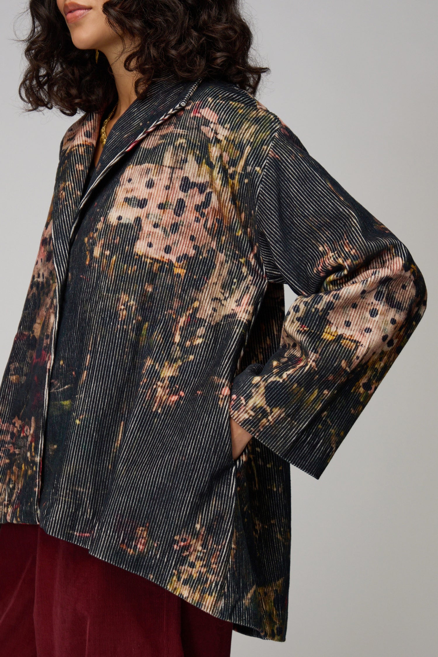 A woman wears the Print Corduroy Jacket, featuring painterly dark green, red, and beige tones, as she stands against a plain background looking slightly to the side.
