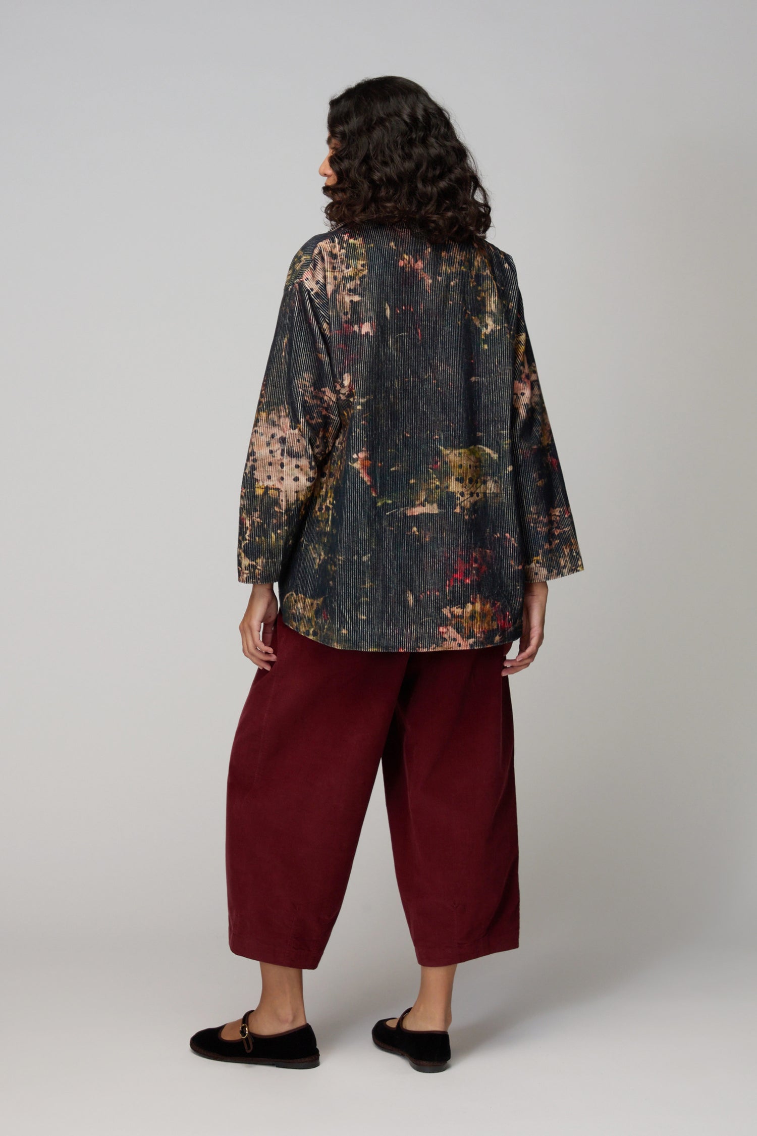 A person stands facing away, wearing a relaxed look with the Print Corduroy Jacket, burgundy wide-leg pants, and black slip-on shoes with white dots against a plain background.