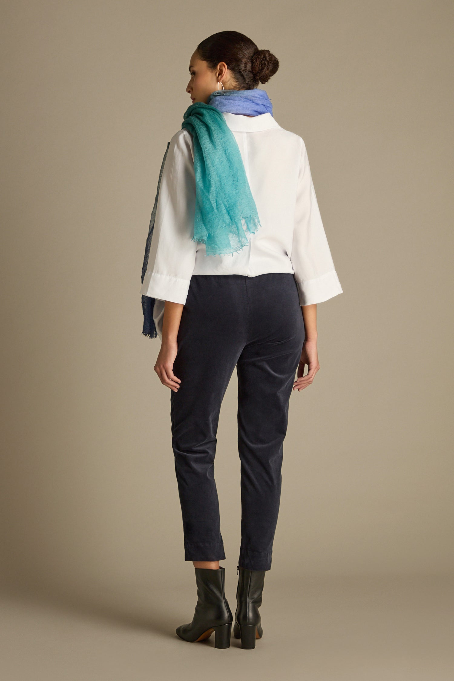 A person dressed in a white shirt, tailored dark pants—highlighted by the elegant silhouette of the Cotton Moleskin Slim Trouser—black boots, and a blue-green scarf stands facing away against a neutral background.
