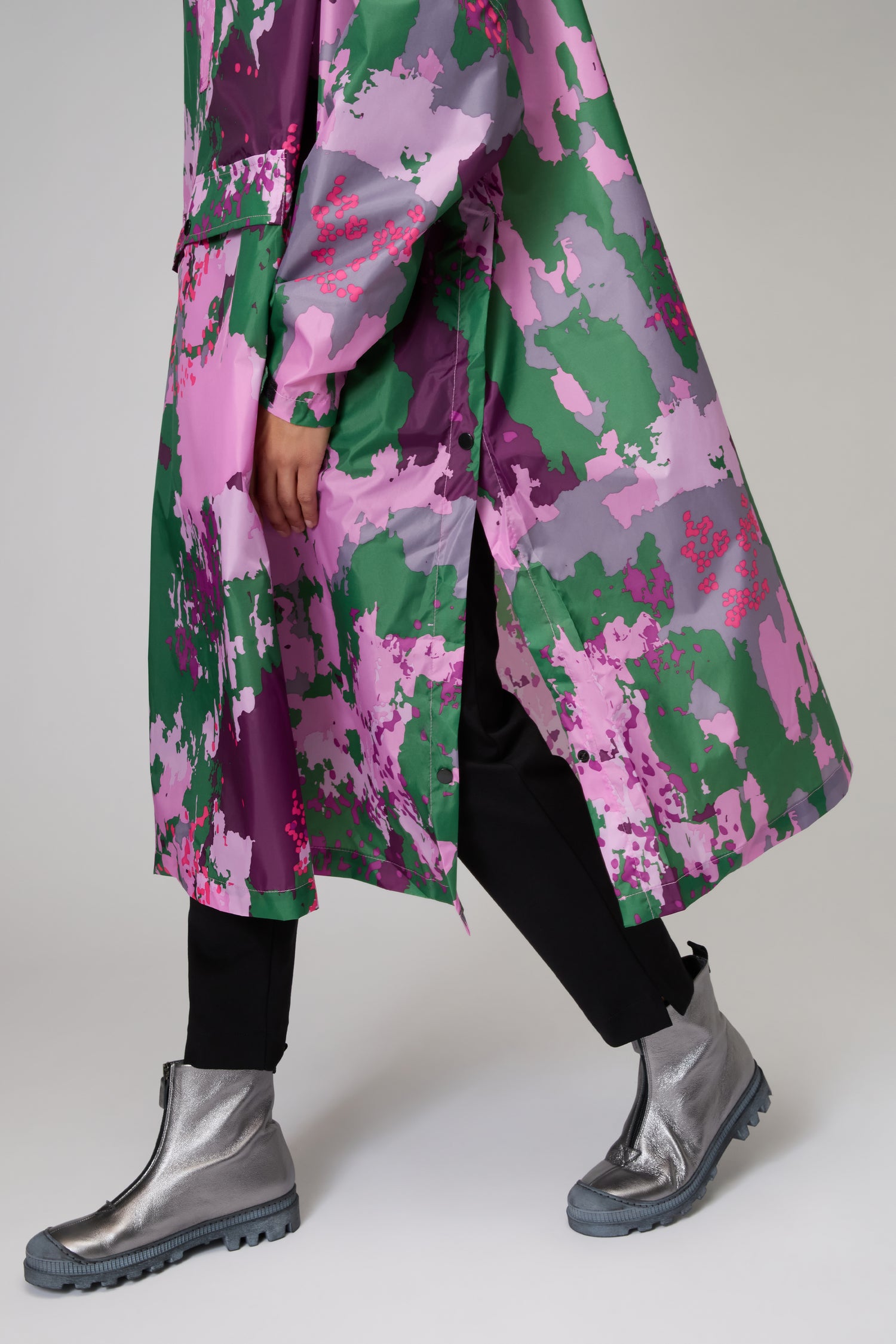 A person wears the Digi Spring Poncho, a long recycled polyester raincoat with a vibrant floral pattern, styled with black pants and silver ankle boots—a modern, sustainable outerwear look.
