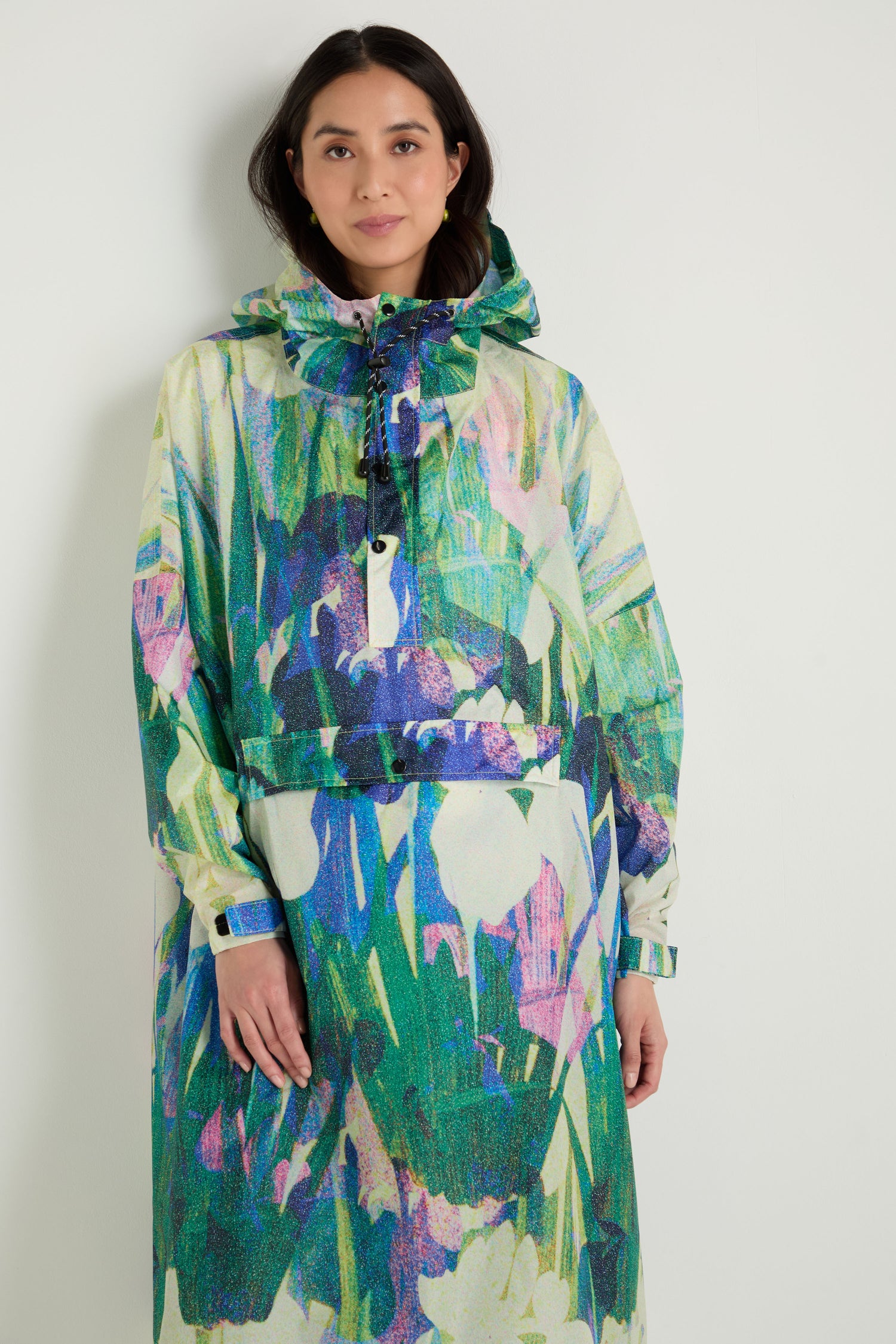 A woman stands against a plain wall wearing the Lily Poncho, a colorful floral rain poncho made from recycled polyester, paired with a matching skirt.