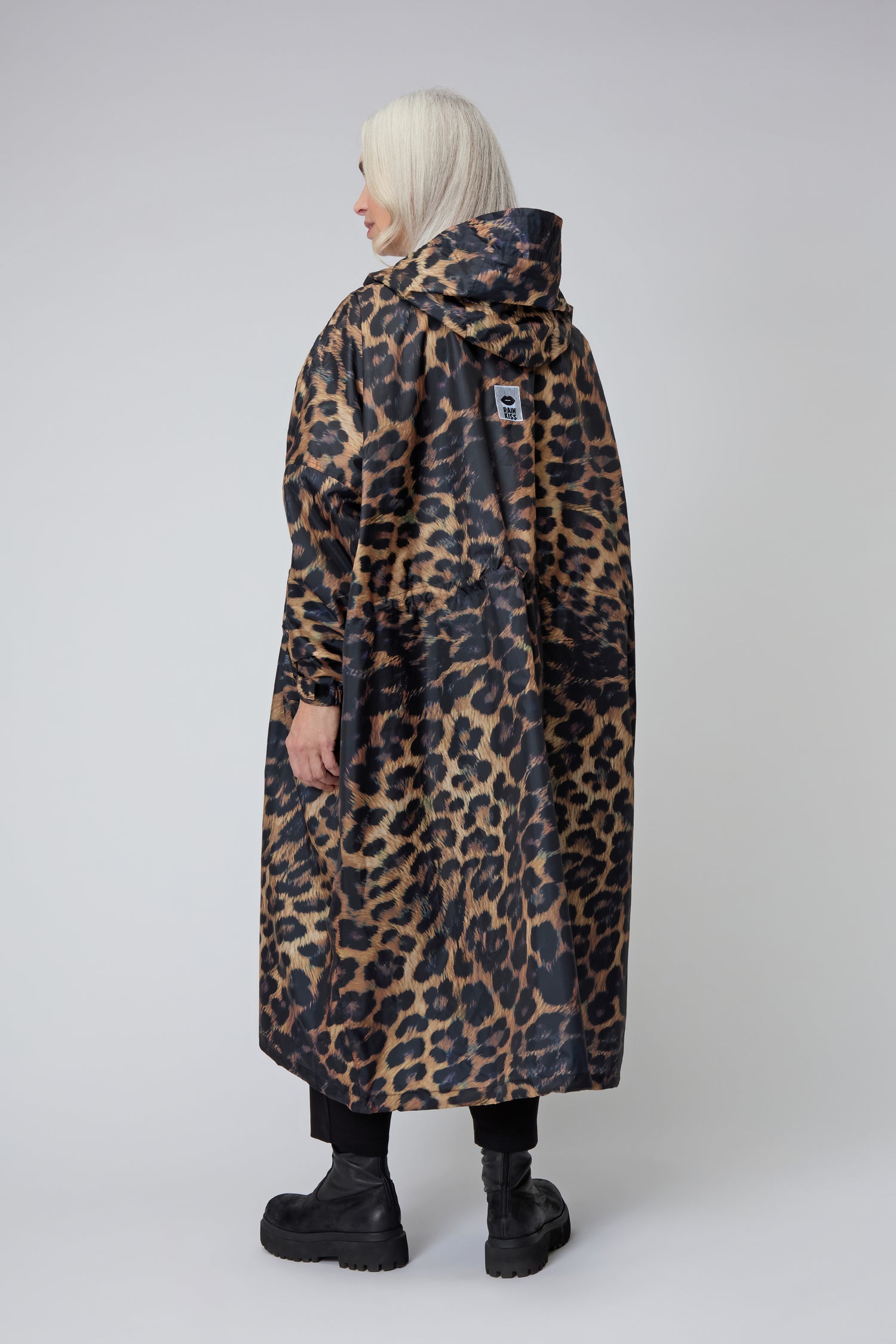 Someone with long gray hair, wearing the Dark Leopard Rain Coat and black boots, stands facing away from the camera against a plain light background. The coat is made using recycled polyester.