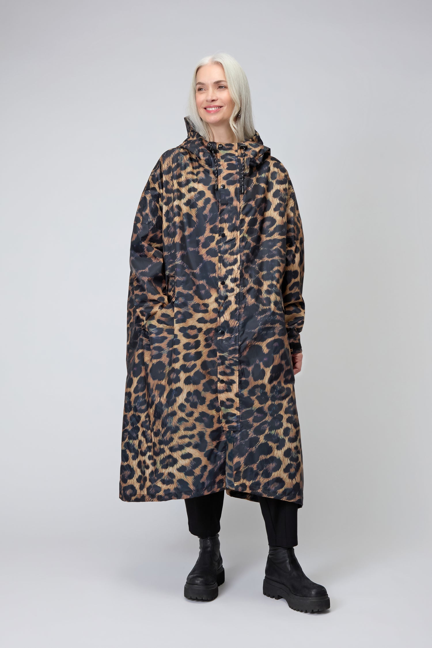A person with long gray hair wears the Dark Leopard Rain Coat, made from recycled polyester, along with black pants and boots, standing against a plain light gray background.