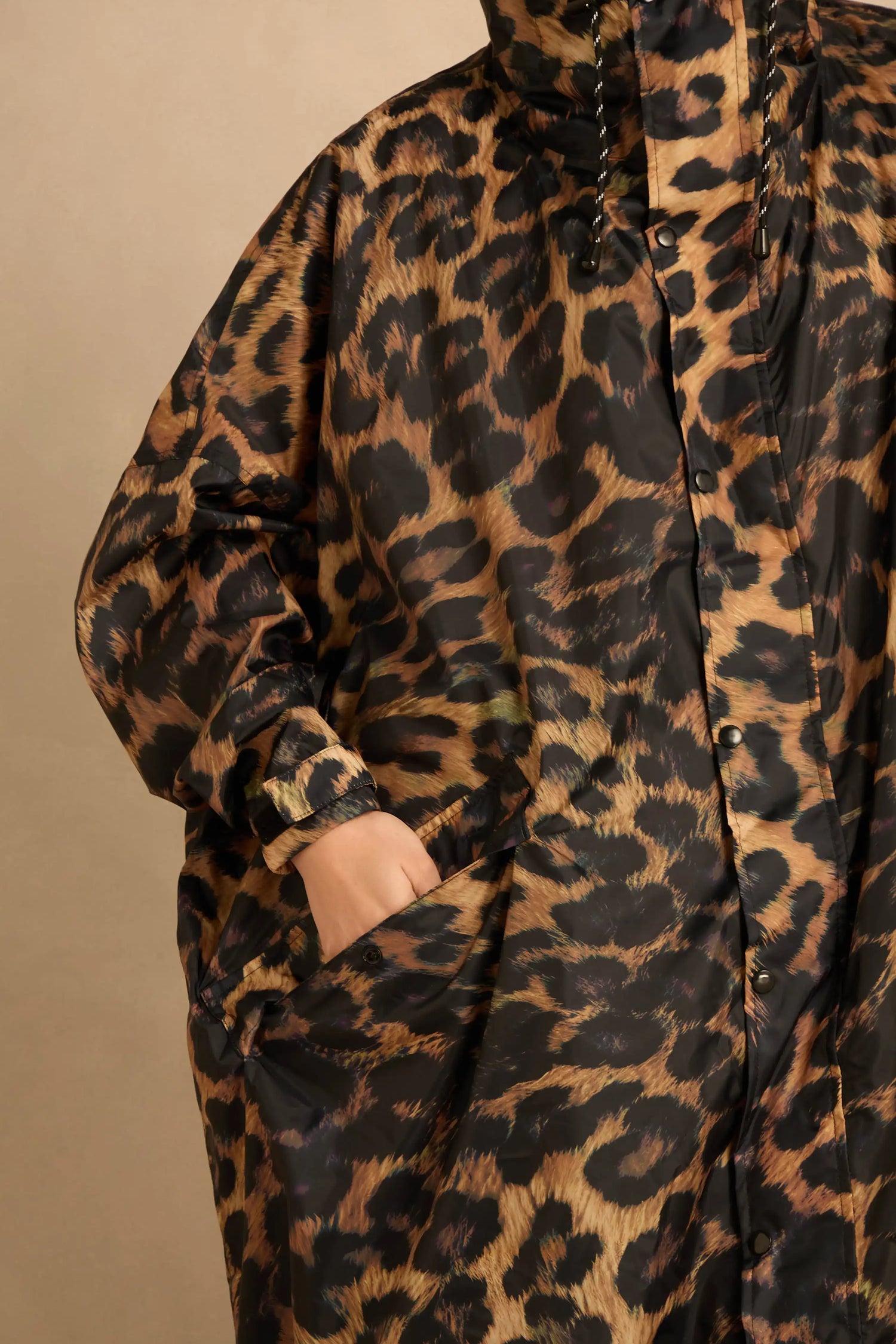 A person wearing an oversized, dark brown and black Dark Leopard Rain Coat with a hood and a hand tucked into the large front pocket.